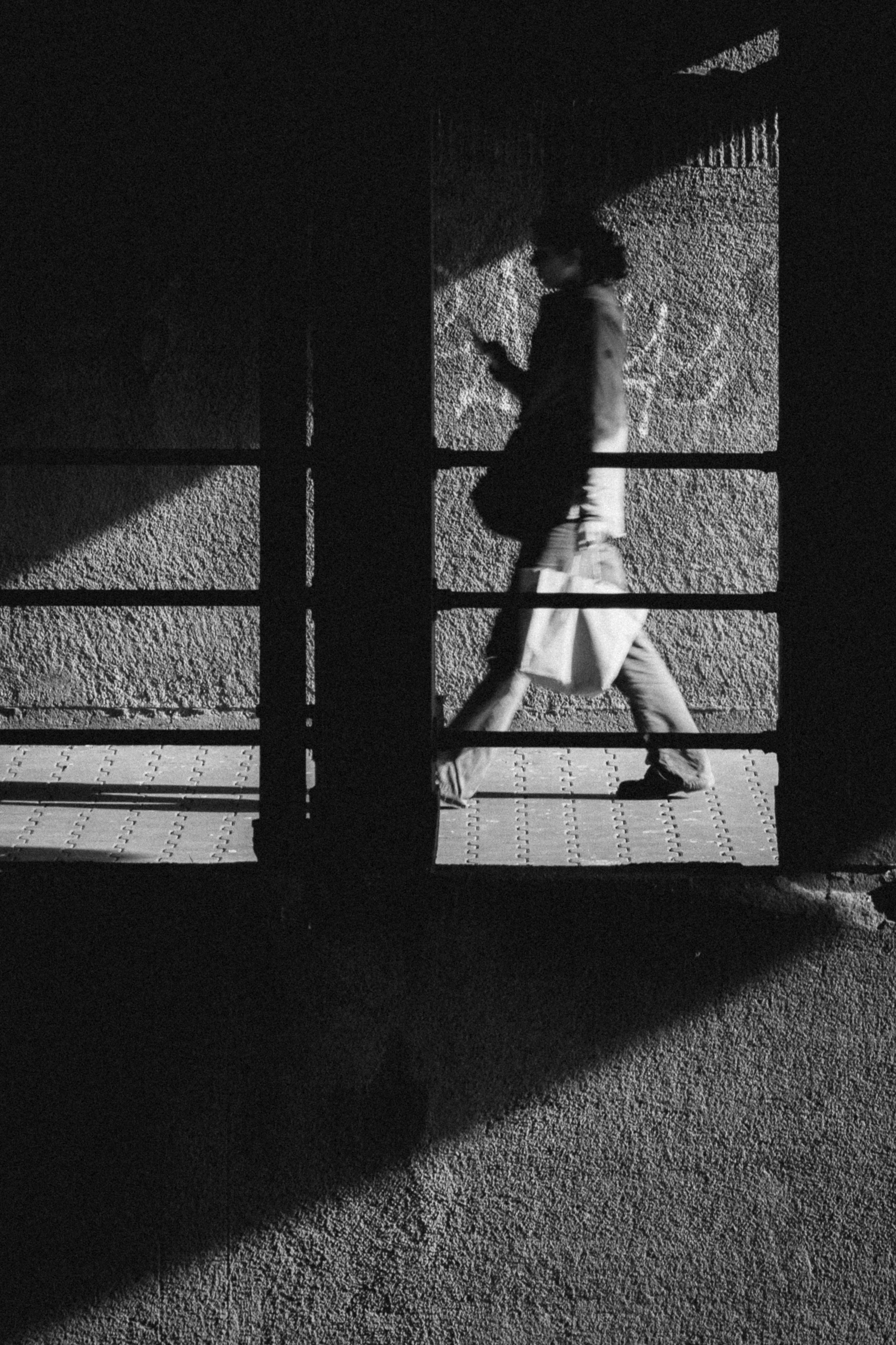 Black and white photograph of a person walking under a bridge in Düsseldorf, Germany. The figure is captured mid-stride, holding a bag, with strong contrast between light and shadow creating a striking urban atmosphere.