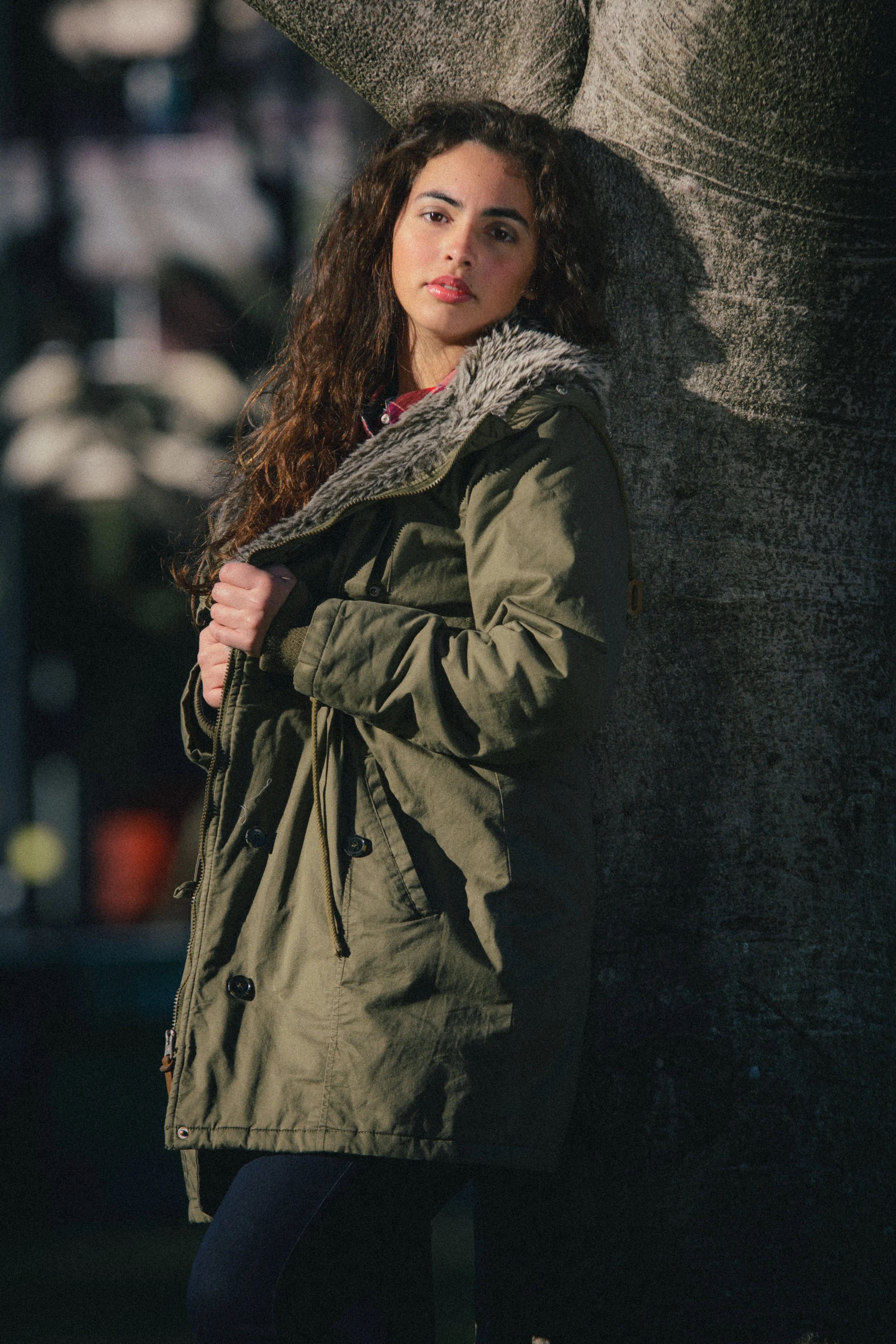 Portrait of Inês Chalupa laying agains a tree, wearing a green coat looking at the camera, grabbing her coat.