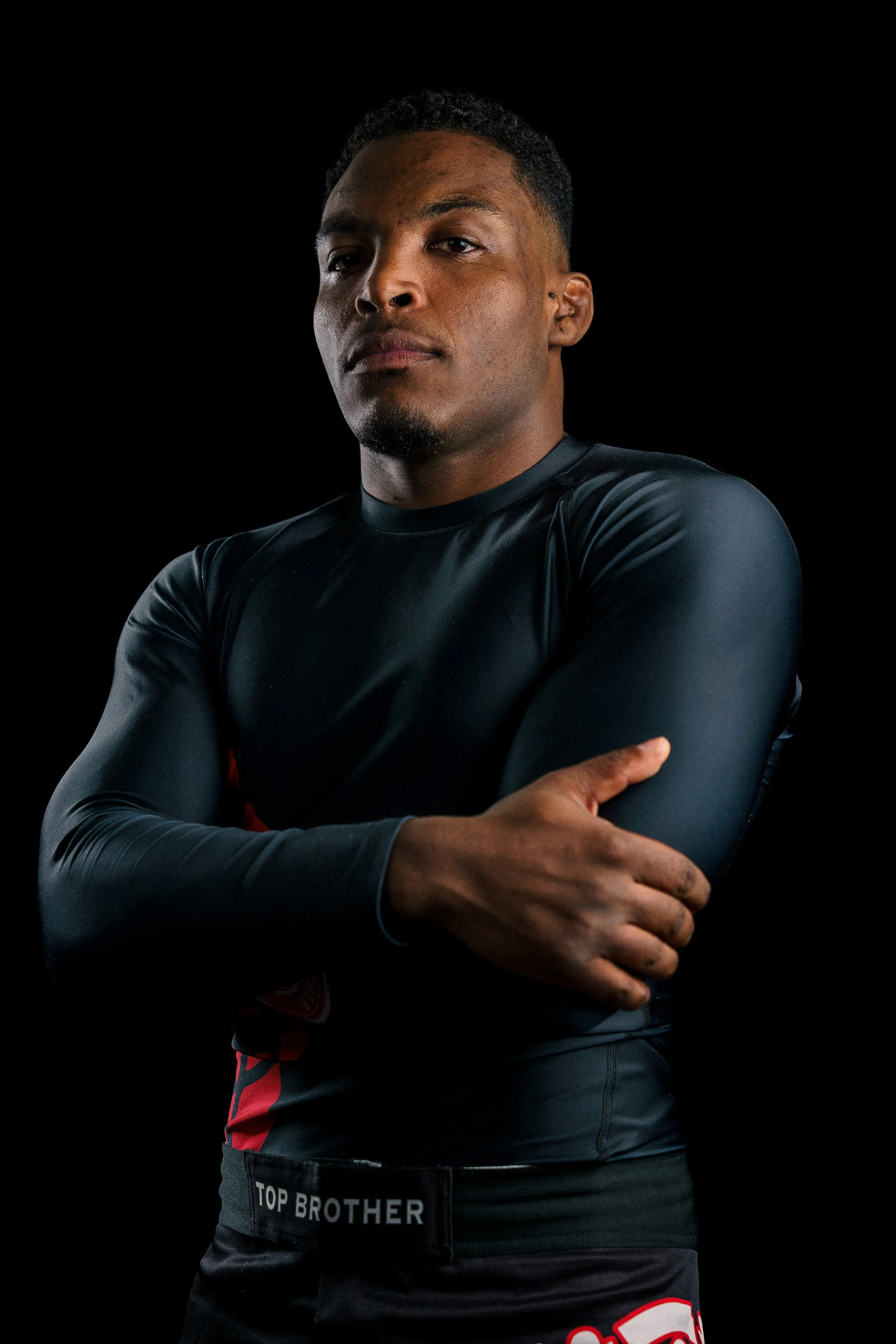Studio portrait of a Brazilian Jiu-Jitsu athlete, Renan Malheiro from Top Brother, in nogi clothes, standing confidently in a training hall, crossed arms.
