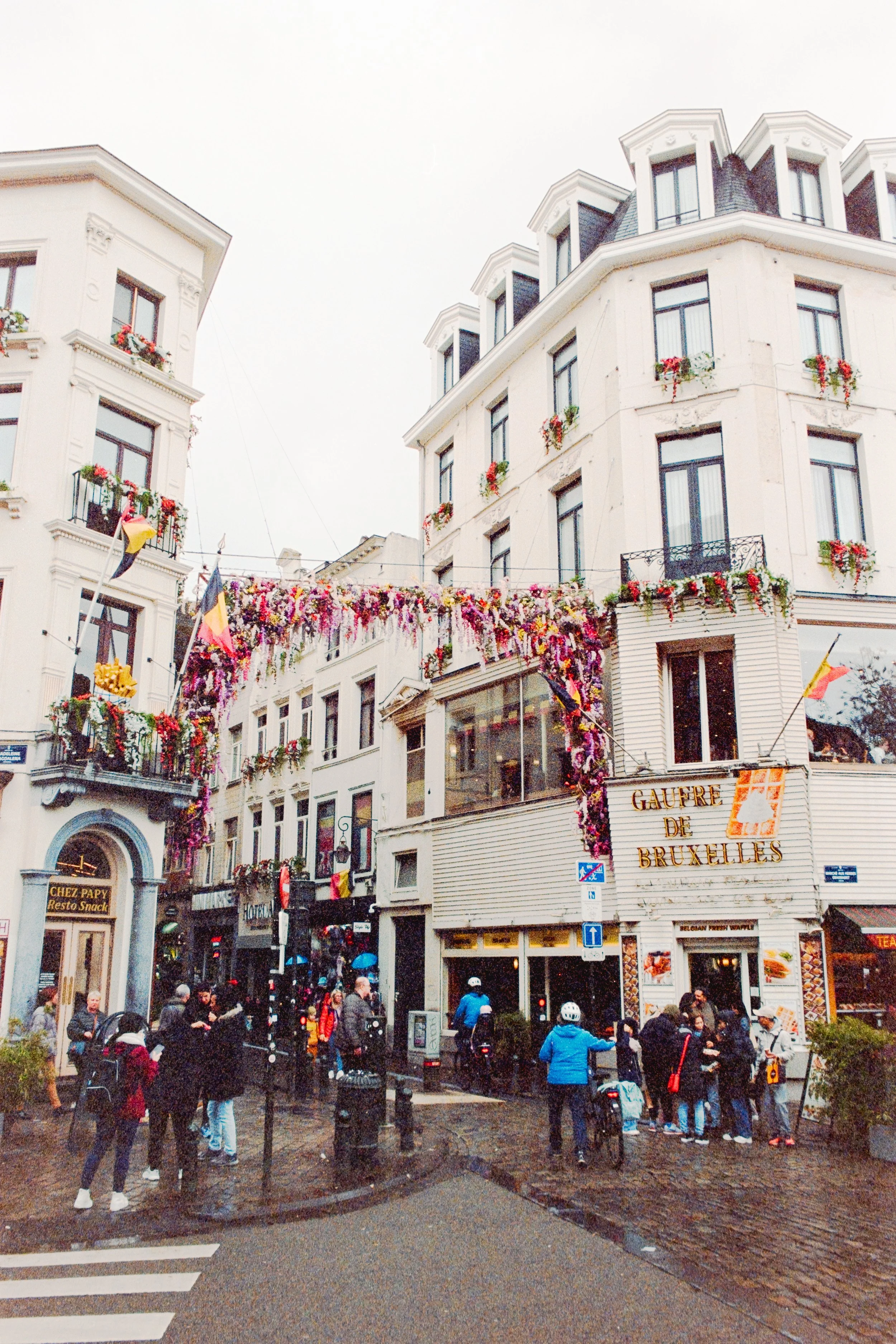 Fine art photograph of the iconic Gaufre de Bruxelles waffle shop in Brussels, Belgium, framed by vibrant floral garlands and cobblestone streets, evoking the charm of European culture and premium urban photography.