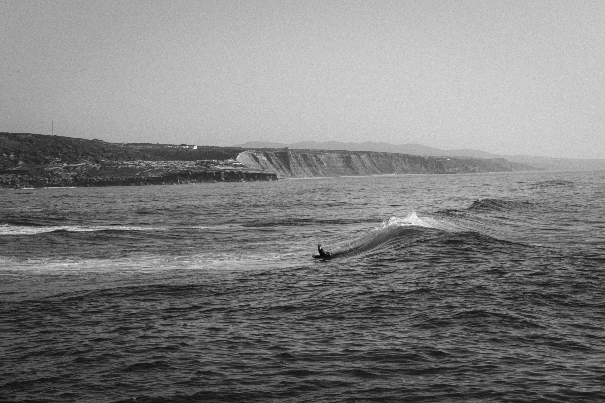 A black-and-white photograph of a surfer riding a wave in the open sea near Ericeira, Portugal. In the background, rugged cliffs stretch along the coastline under a hazy sky.