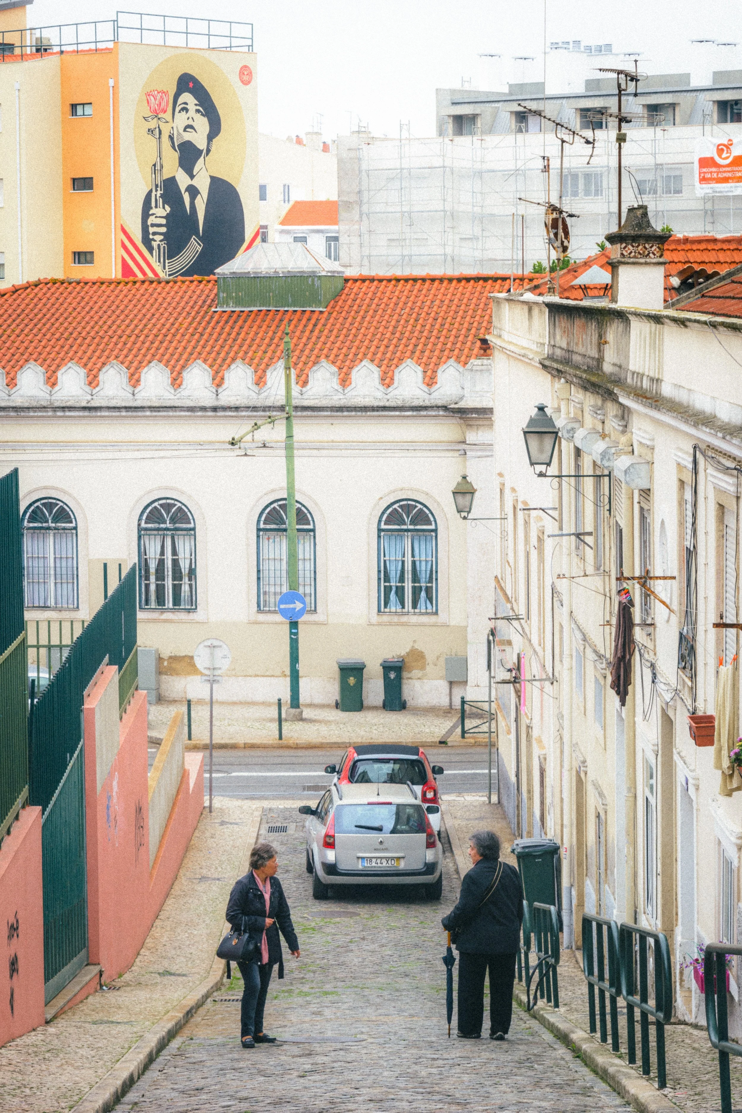 A cobblestone street in Lisbon, Portugal, where two women are engaged in conversation. The background showcases a vibrant mural symbolizing the 25th of April Revolution, featuring a figure holding a carnation and an iconic rifle. Traditional Portugue