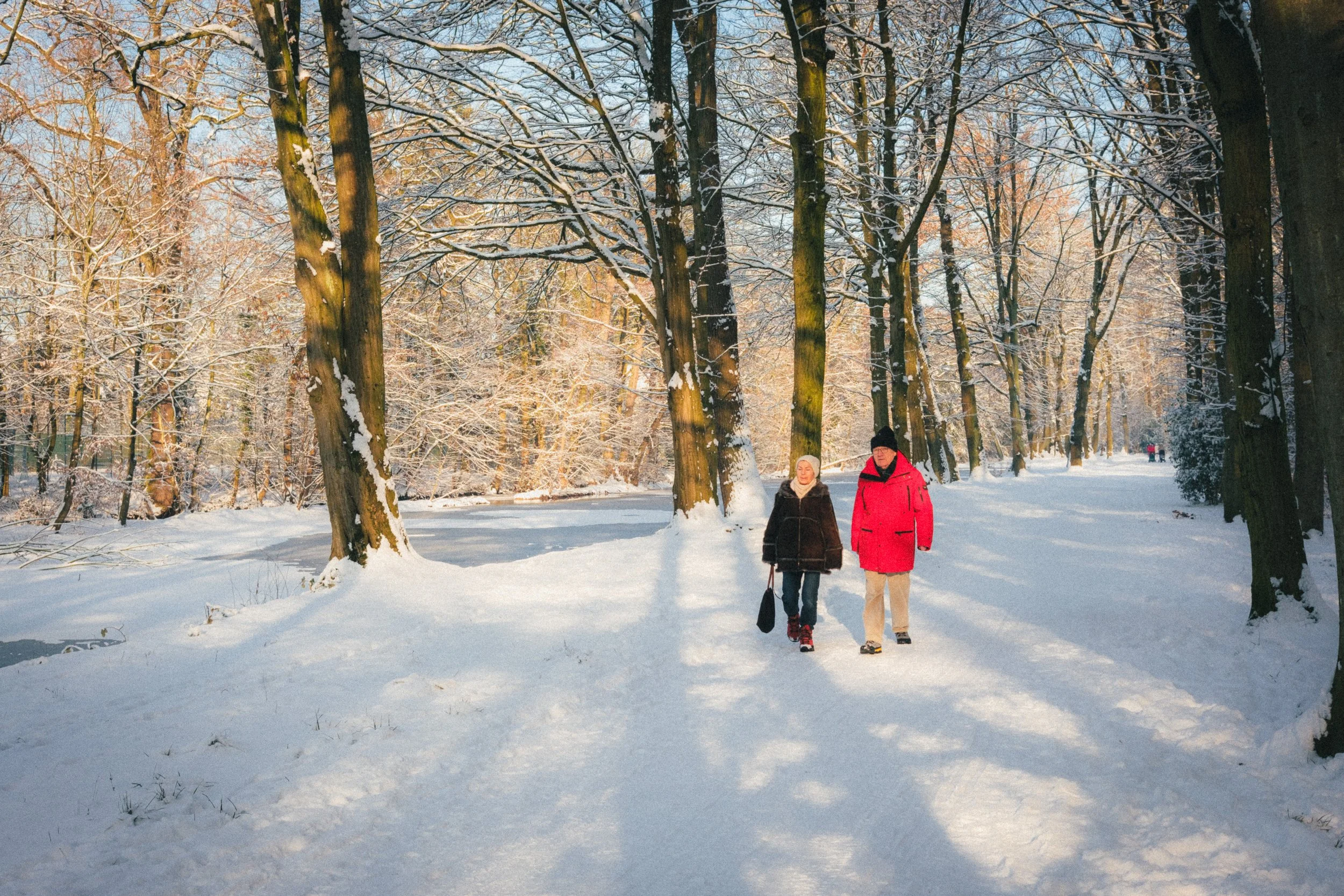 Fine art travel photography of two people walking on a snowy path surrounded by trees in Schloss Benrath Park, Düsseldorf, Germany, with winter sunlight casting long shadows.