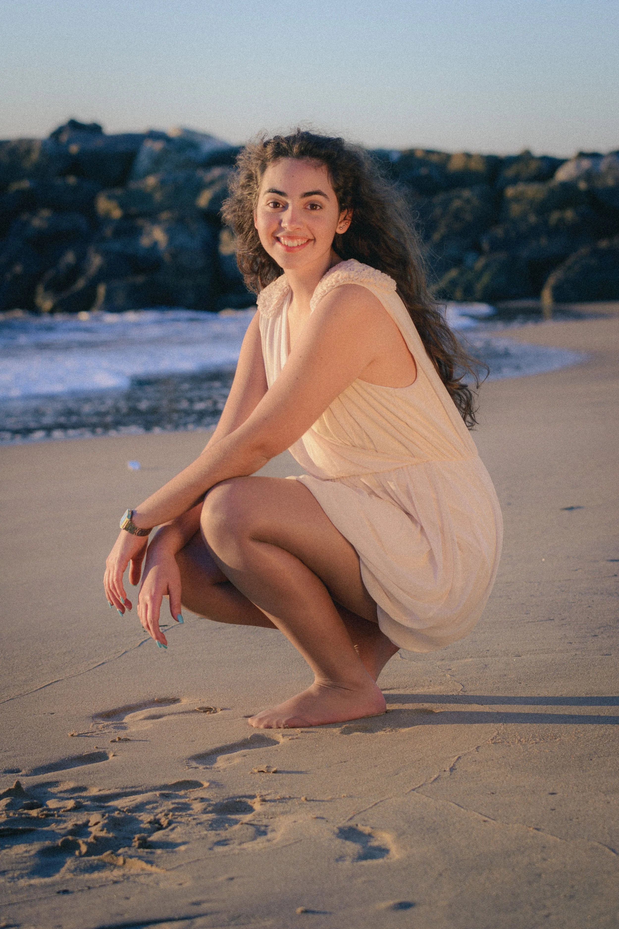 Portrait of Inês Chalupa in a beach in Portugal in Golden hour, wearing a white dress smiling at the camera, standing in the sand with the blue water behind her.