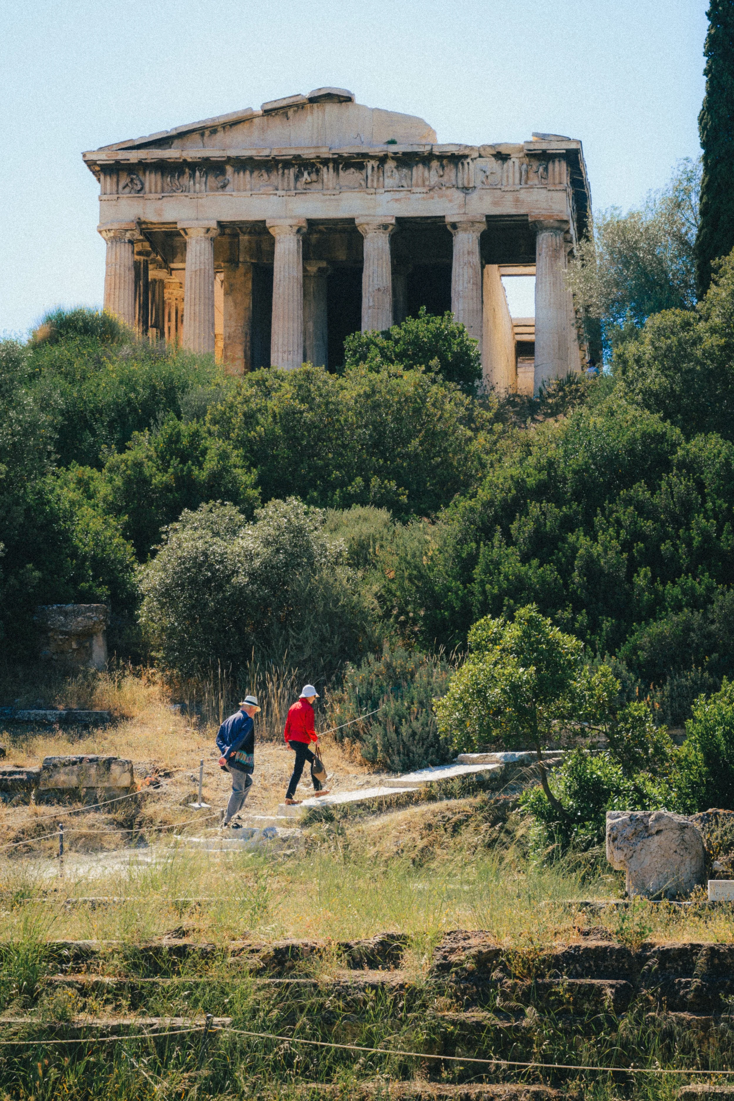 A photograph taken in Athens, Greece, featuring two people walking on a path surrounded by lush greenery. Above them stands an ancient Greek temple, its classical columns and structure partially weathered, adding a sense of historical grandeur. The s