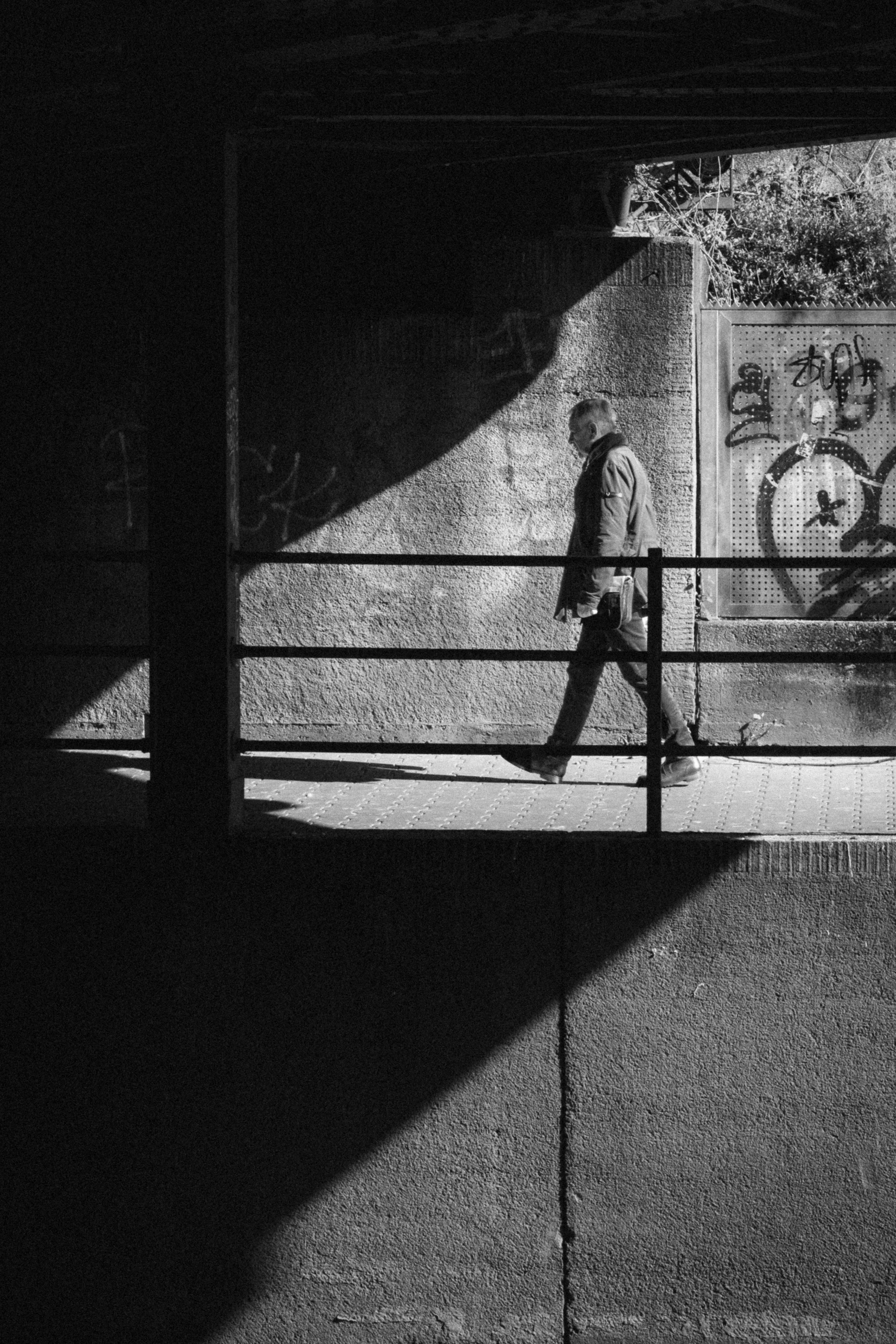Black and white photograph of a man walking under a bridge in Düsseldorf, Germany. The composition uses dramatic light and shadow to highlight the figure against graffiti-covered walls, evoking an urban and contemplative mood.