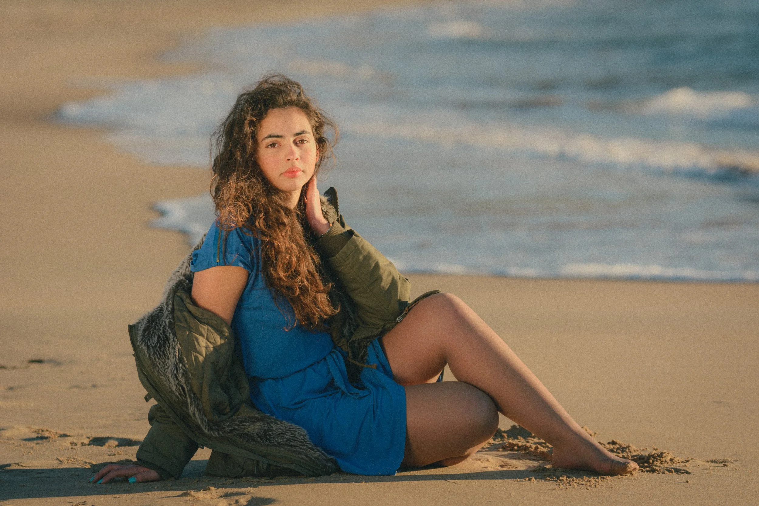 Portrait of Inês Chalupa in a beach in Portugal in Golden hour, wearing a blue dress and a green coat looking at the camera, sitting in the sand with the blue water behind her.
