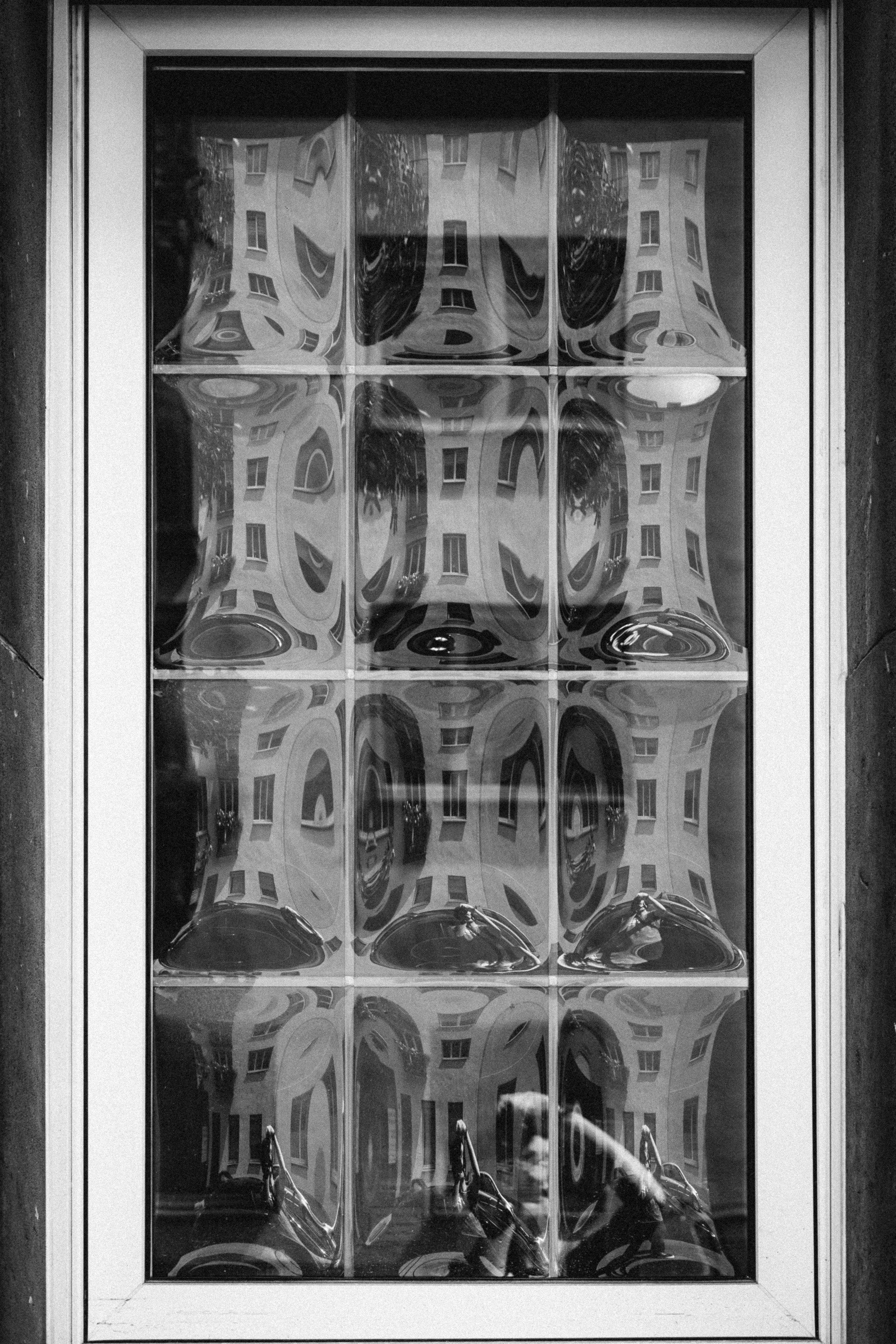 A monochrome photograph of a distorted reflection captured through a textured glass window. The warped glass panes create abstract patterns of the building opposite, with its windows and facade bending and curving in surreal forms. The lower section 