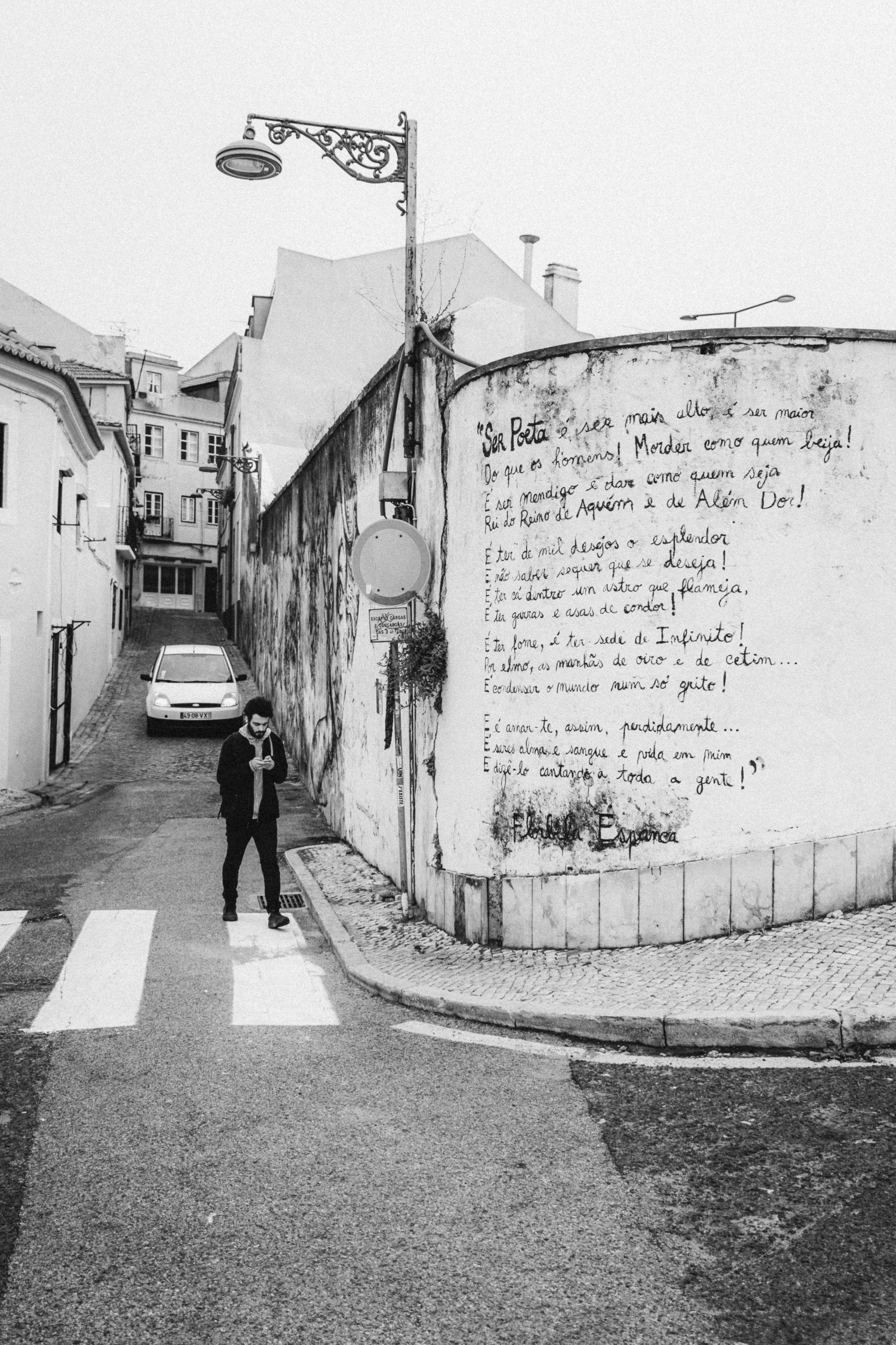 A street scene in Lisbon, Portugal, featuring a man crossing a zebra crossing while looking at his phone. On the right, a weathered wall displays a poem by Florbela Espanca, adding a layer of artistic and cultural depth to the urban setting. The blac