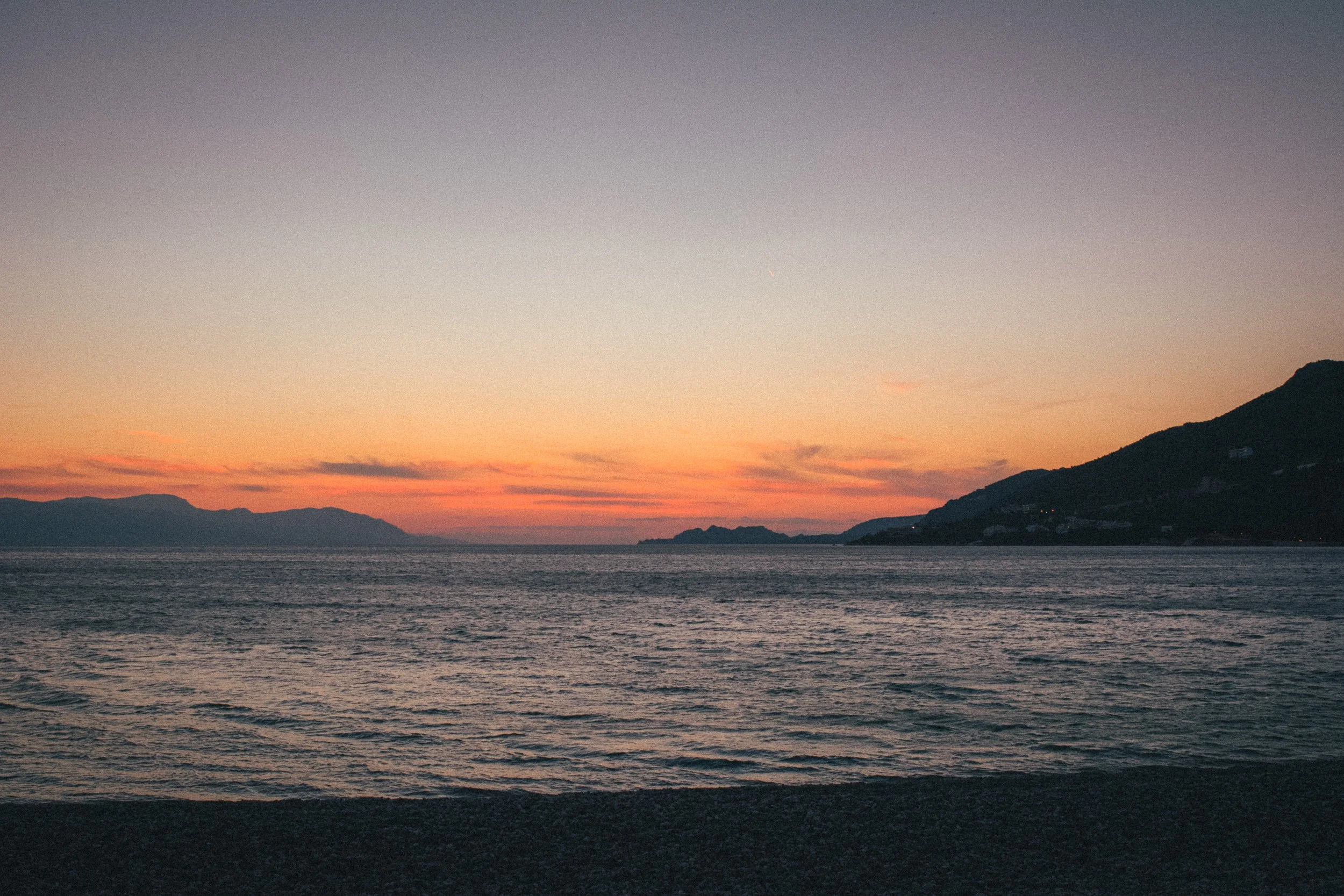 Fine art landscape photograph of a sunset in Greece. In the foreground the sand, followed by the cold sea and the orange sun in the horizon with the orange/blue sky on top.