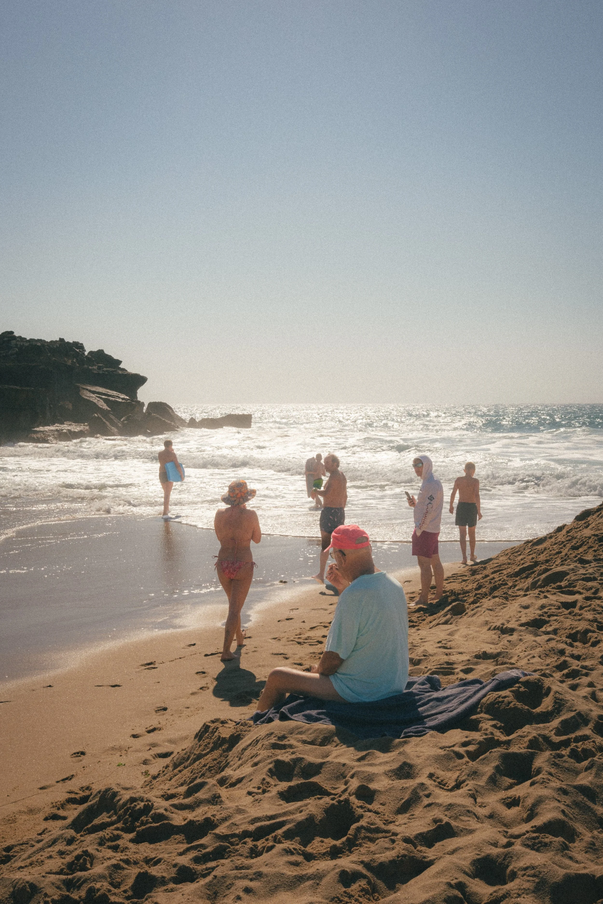 Fine art travel photography of a sunny beach in Ericeira, Portugal, with people enjoying the ocean waves and relaxing on the sandy shore near rocky cliffs.