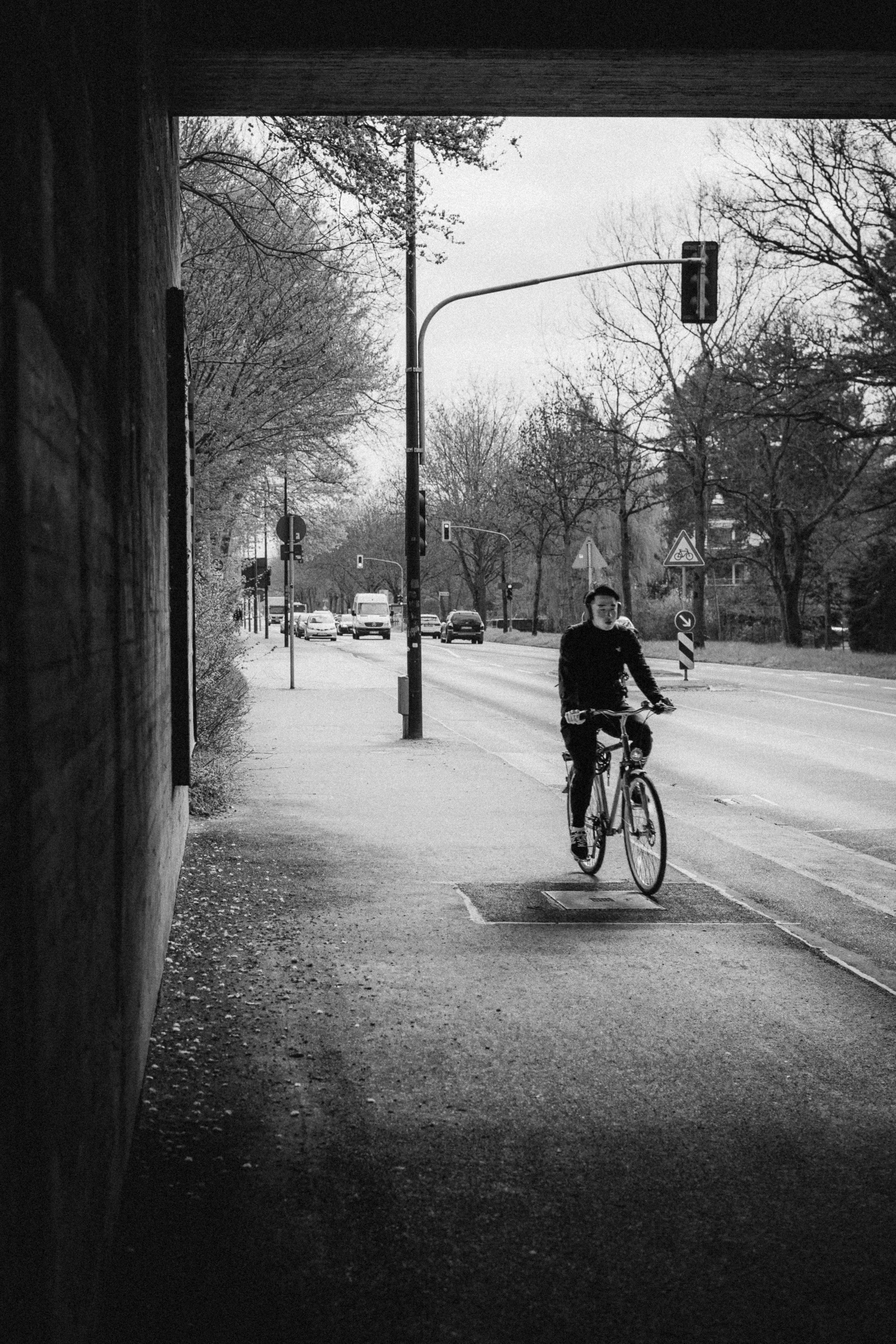 Black and white street photograph of a cyclist riding through an underpass in Düsseldorf, Germany. The composition frames the rider against a busy urban street with traffic lights and tree-lined sidewalks, blending motion and structure.