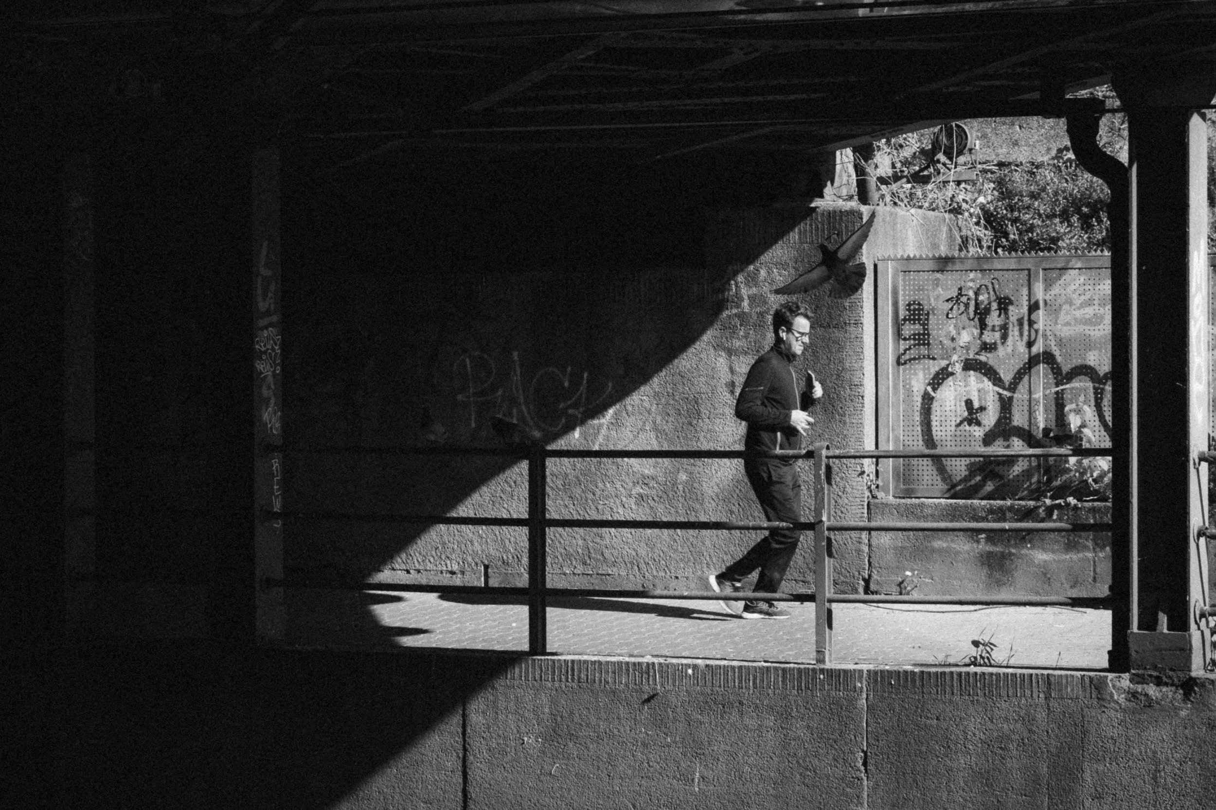 Black and white photograph of a jogger running under a bridge in Düsseldorf, Germany. The interplay of light and shadow accentuates the motion of the runner against a backdrop of graffiti-covered walls, creating a dynamic urban scene.