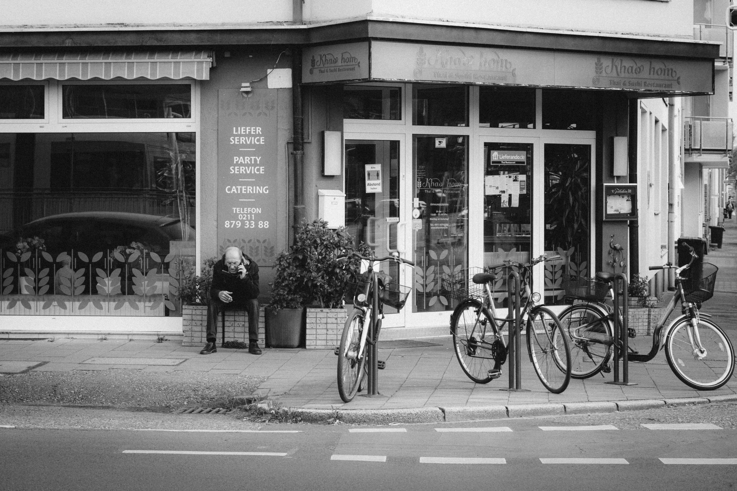 Black and white street photograph taken in Düsseldorf, capturing a moment outside a Thai and sushi restaurant. A man sits on a bench, engrossed in a phone call, while bicycles are parked in the foreground, adding to the urban charm and everyday life 