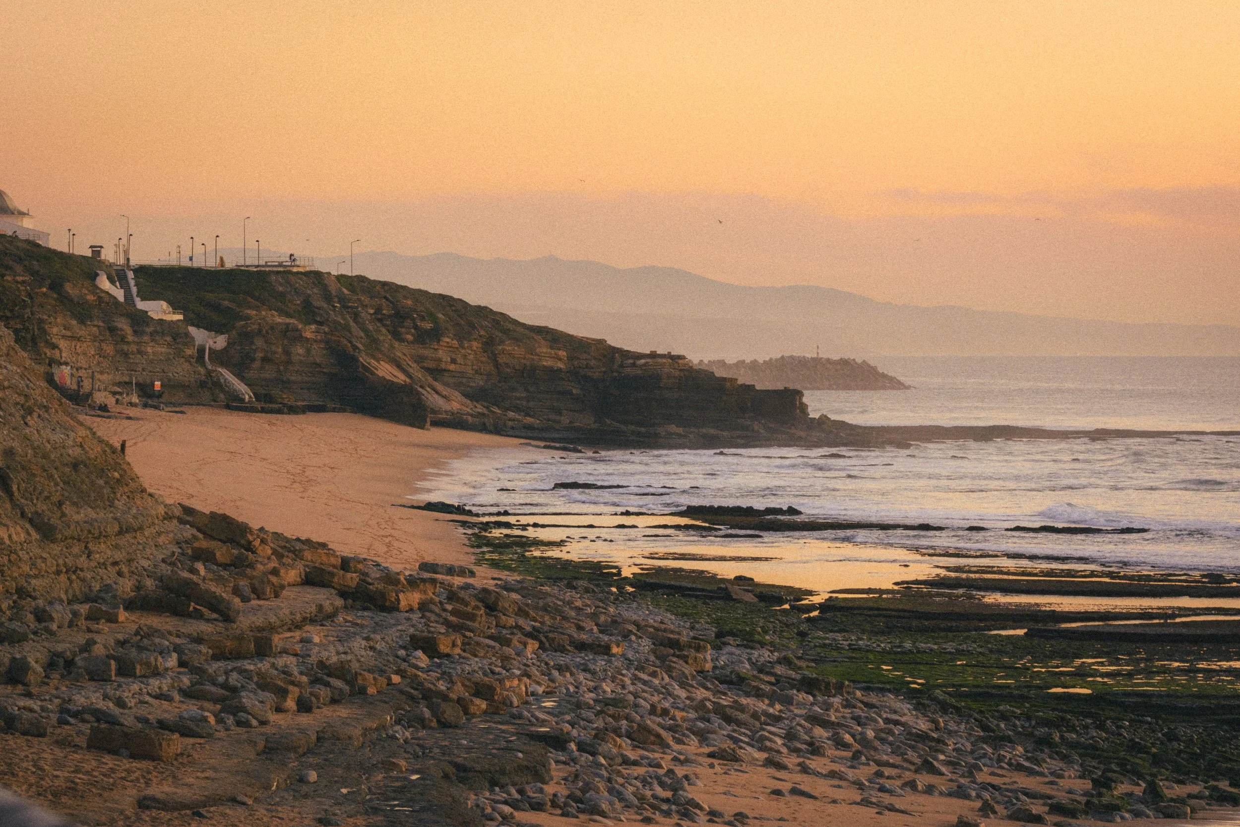 Fine art landscape photograph of a beach in Ericeira, Portugal. Several layers are visible rocks, then water, then hill and finally the orange/yellow sky in a golden hour.