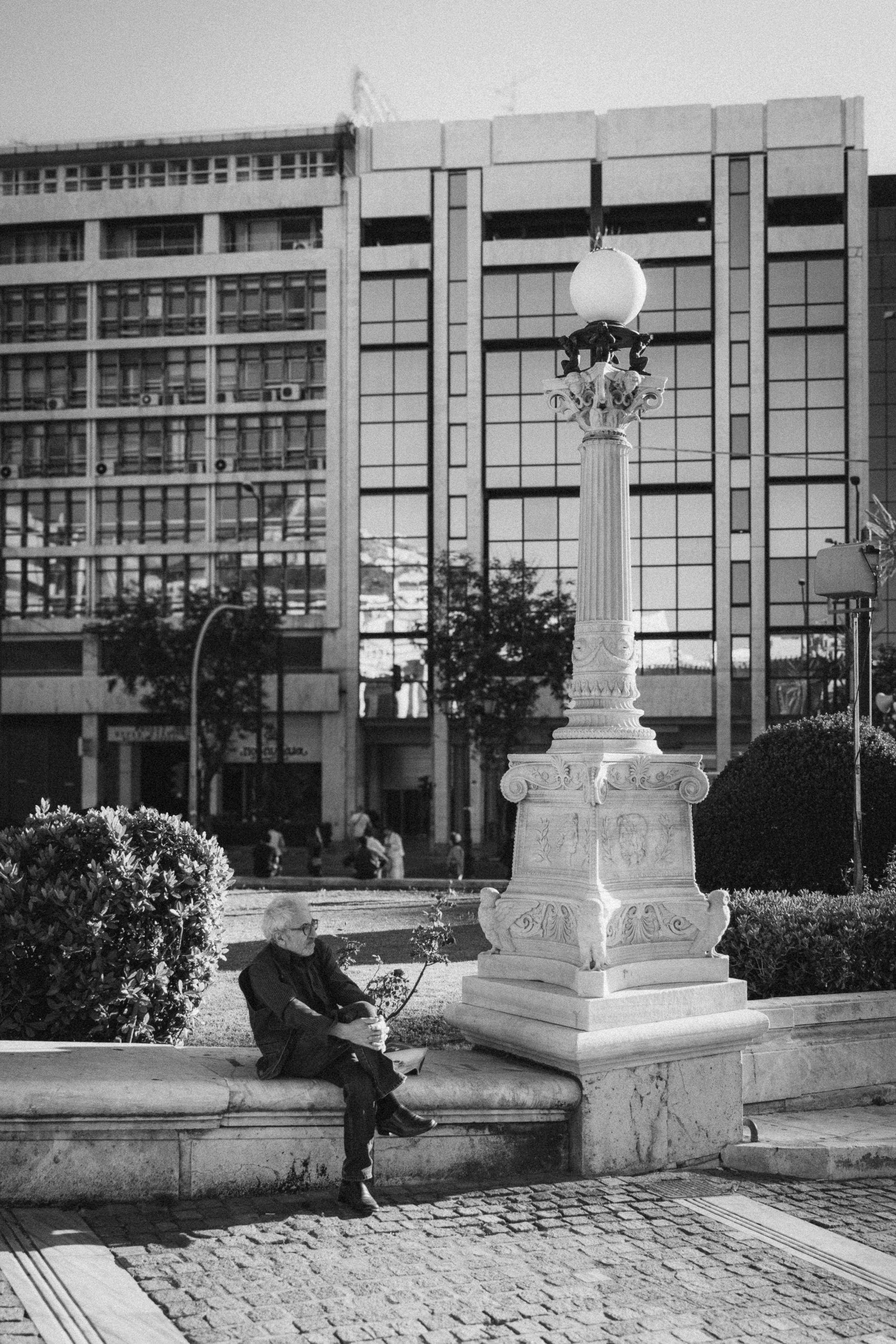 A black-and-white photograph captured in Athens, Greece, showcasing an elderly man seated on a marble bench near an ornate lamp post. The man appears relaxed, gazing off into the distance, with modern glass-fronted buildings in the background. The co