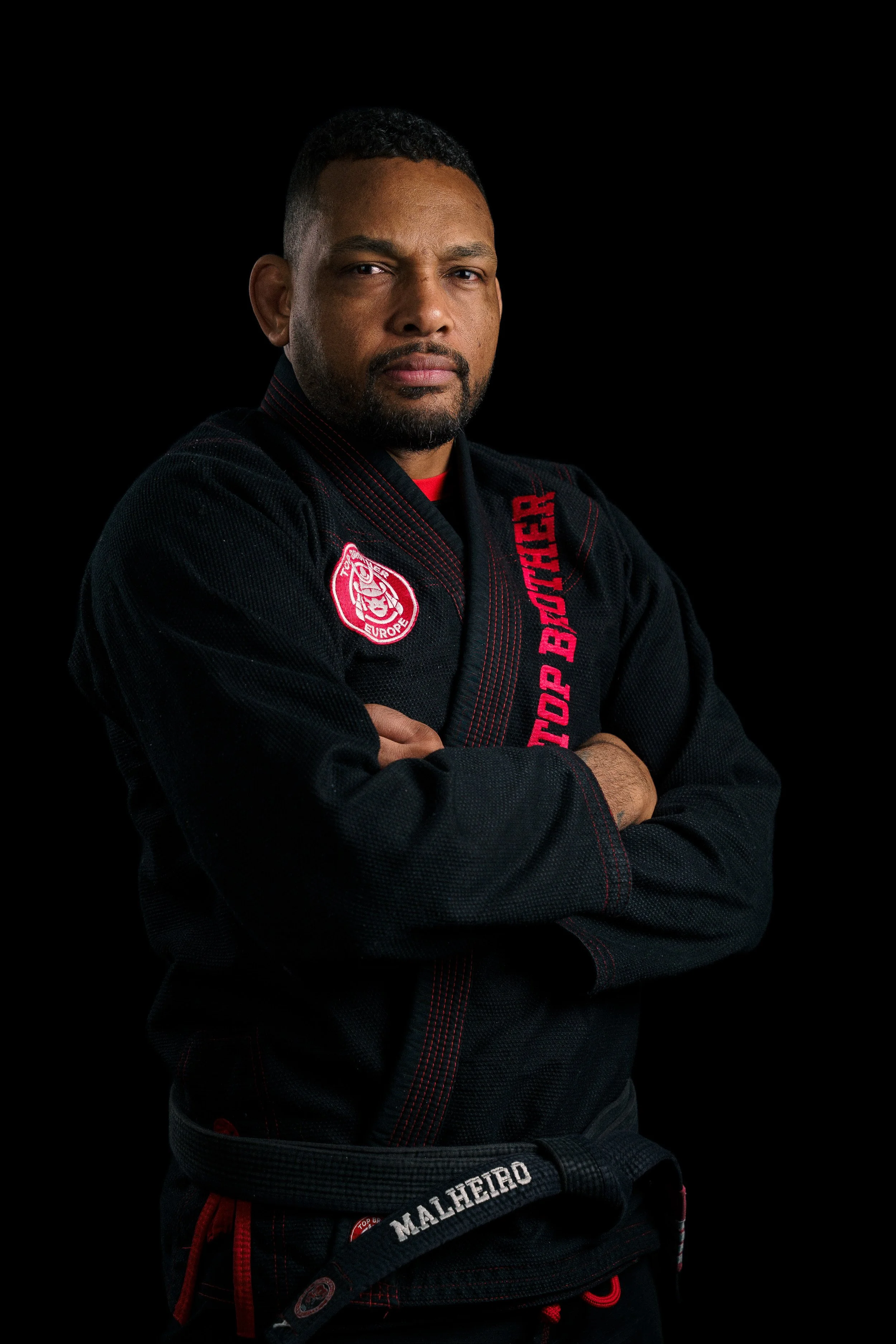 Studio portrait of a Brazilian Jiu-Jitsu athlete, Alex Malheiro from Top Brother, in a black gi, standing confidently in a training hall.