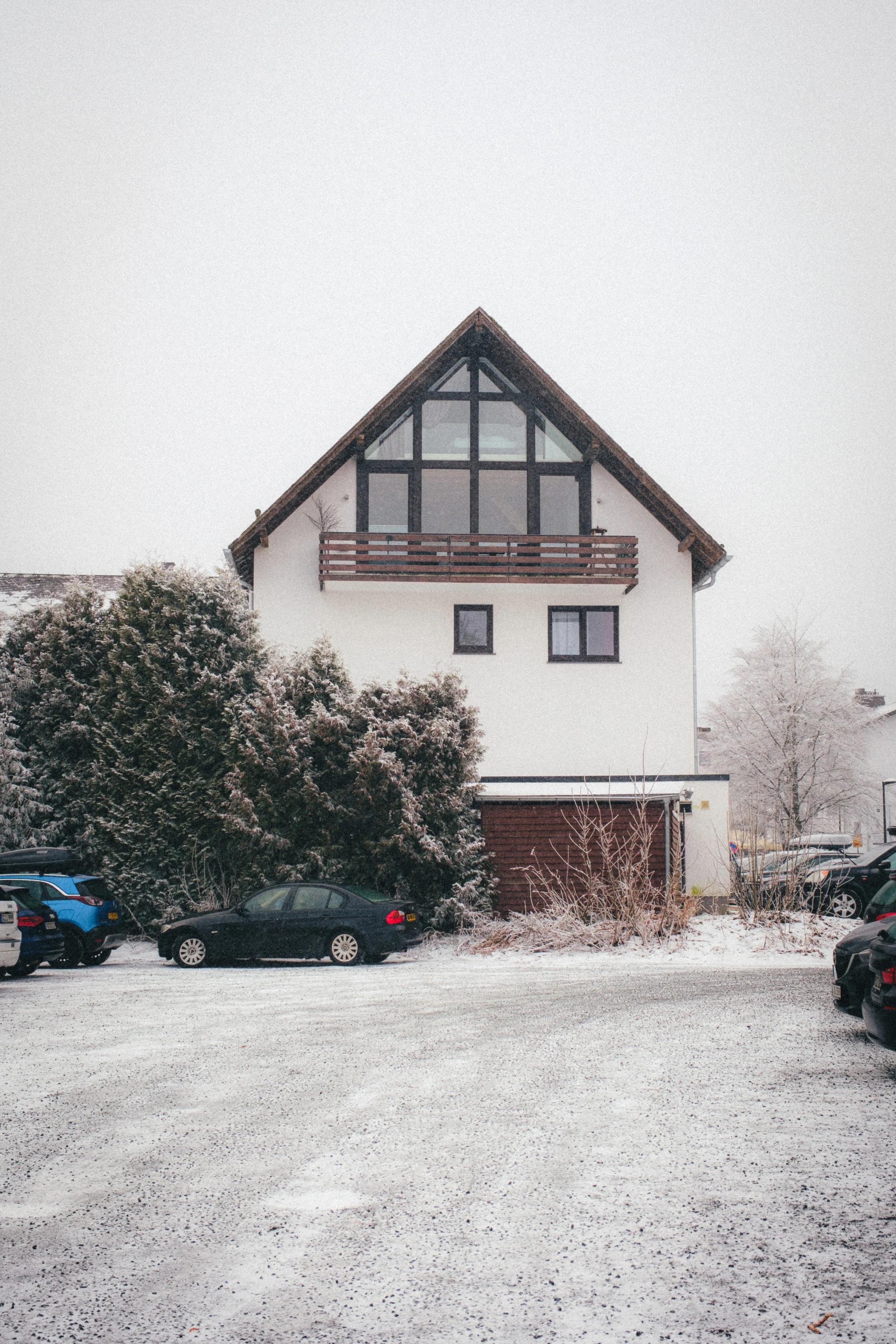 Snow-covered house in Winterberg, Germany, featuring a steep gable roof with large glass windows and a wooden balcony. The scene includes parked cars and frosted trees, capturing the serene atmosphere of a winter day.