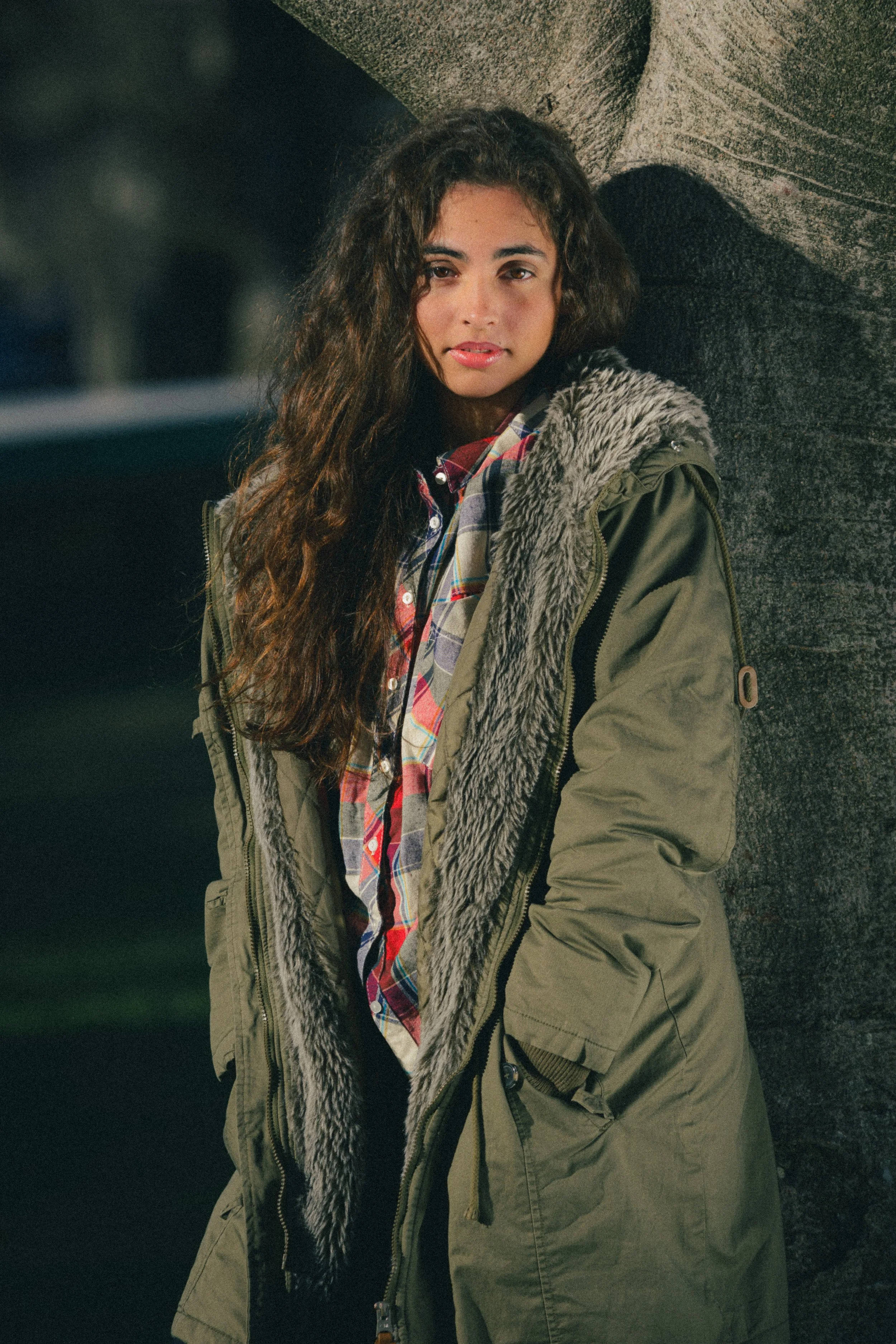 Portrait of Inês Chalupa laying agains a tree, wearing a green coat looking at the camera, with her hands in her pockets..