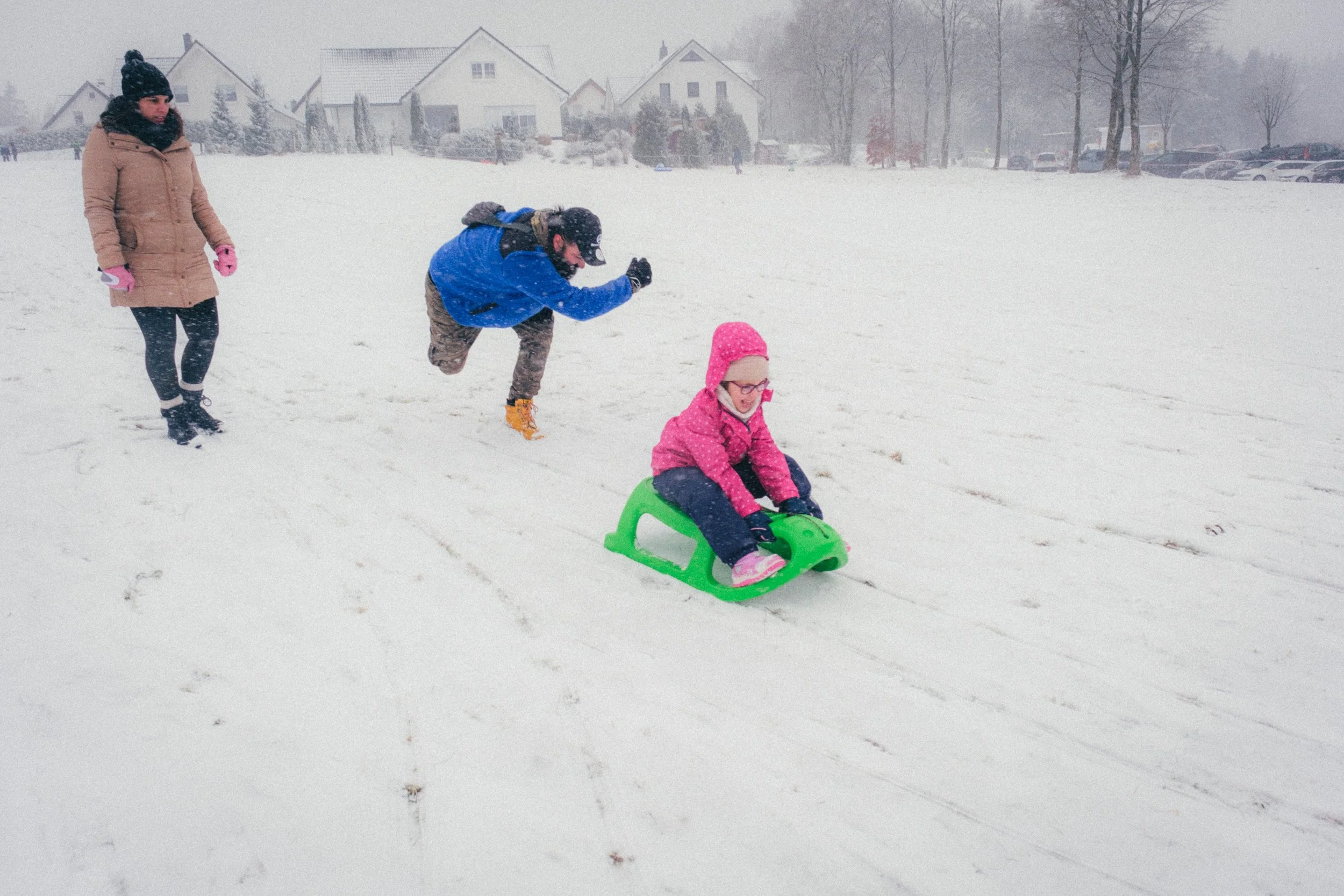 Fine art travel photography of a winter scene in Winterberg, Germany, featuring a child sledding on a green sled, pushed by her father, with snow-covered houses and trees in the background.