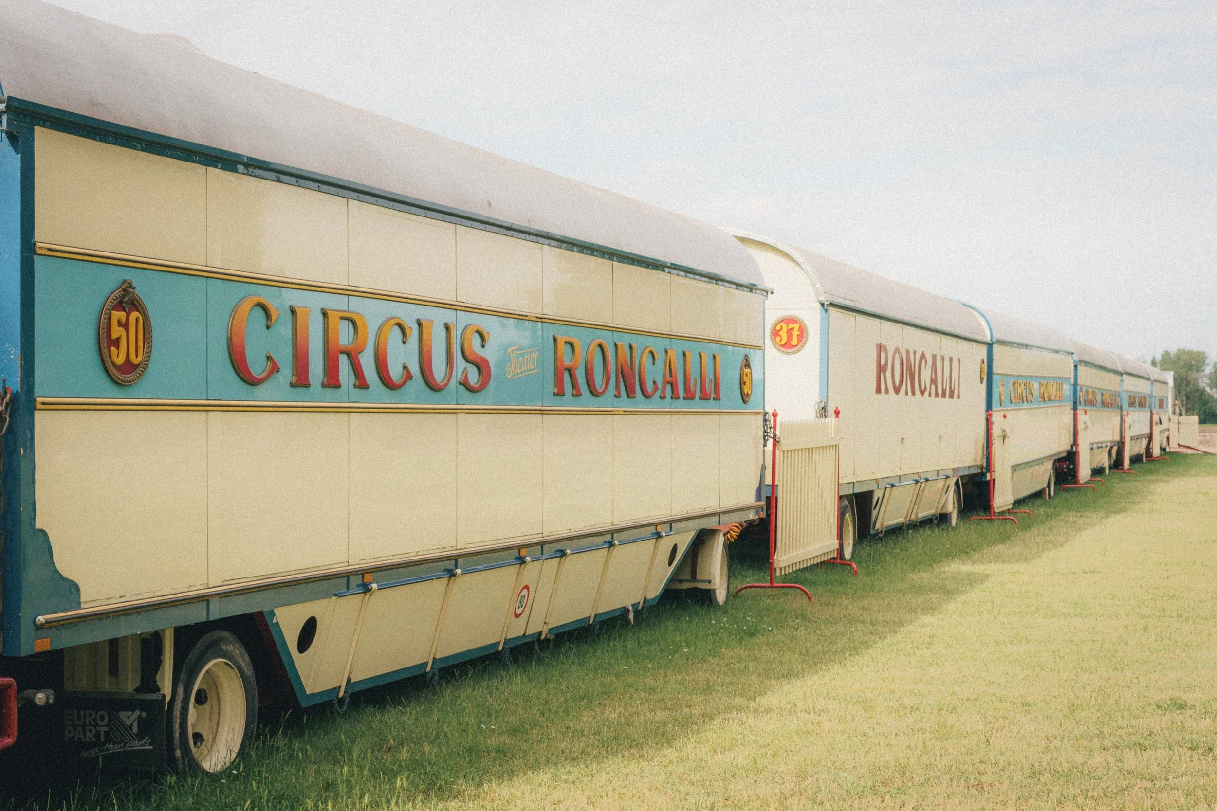 A row of vintage Circus Roncalli trailers painted in cream and blue with red lettering, parked on a green grass field under a clear sky in Düsseldorf. The trailers are numbered, with one showing the number 50 prominently.