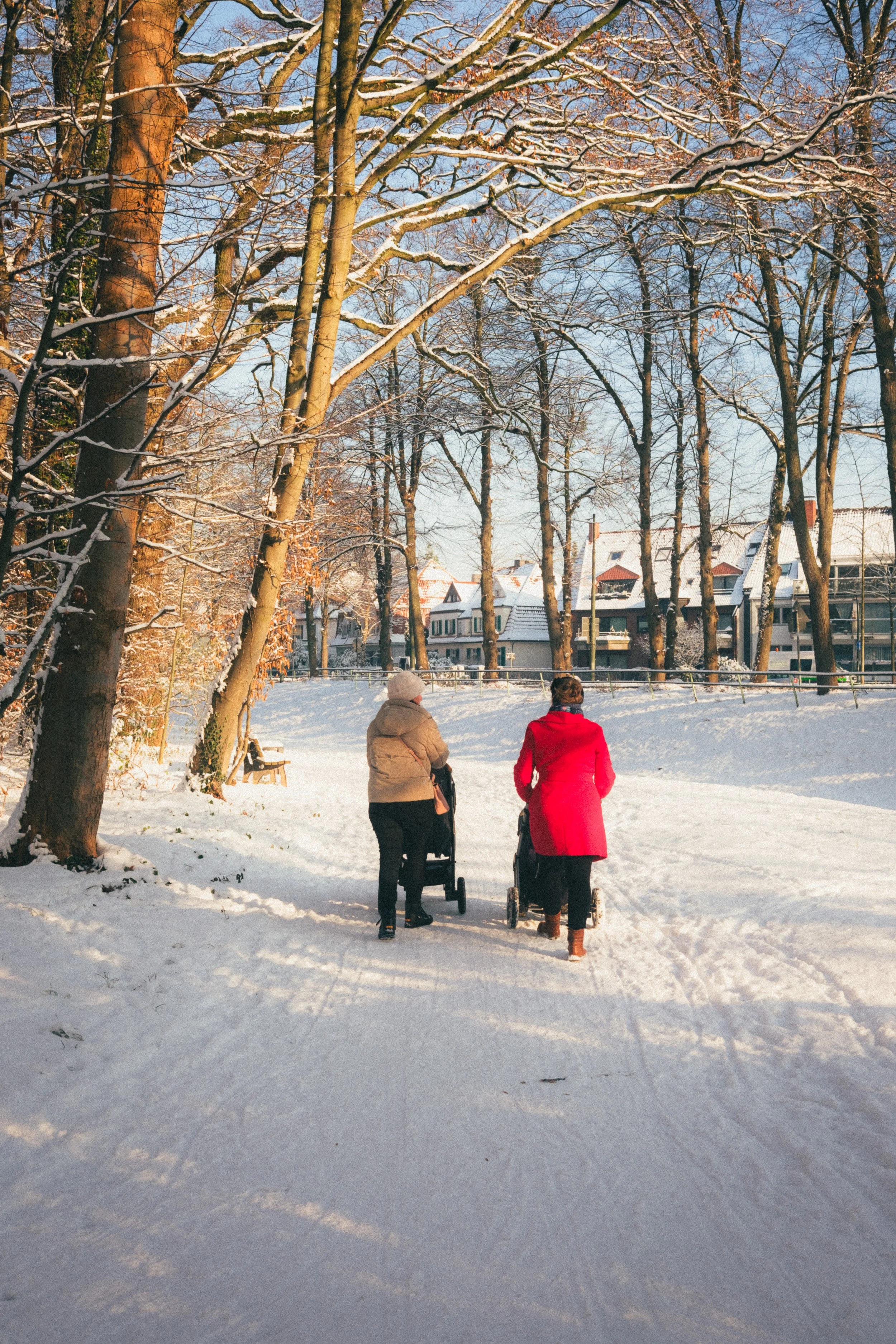 Fine art travel photography of two women walking with strollers on a snowy path in Schloss Benrath Park, Düsseldorf, Germany, with houses visible in the background.