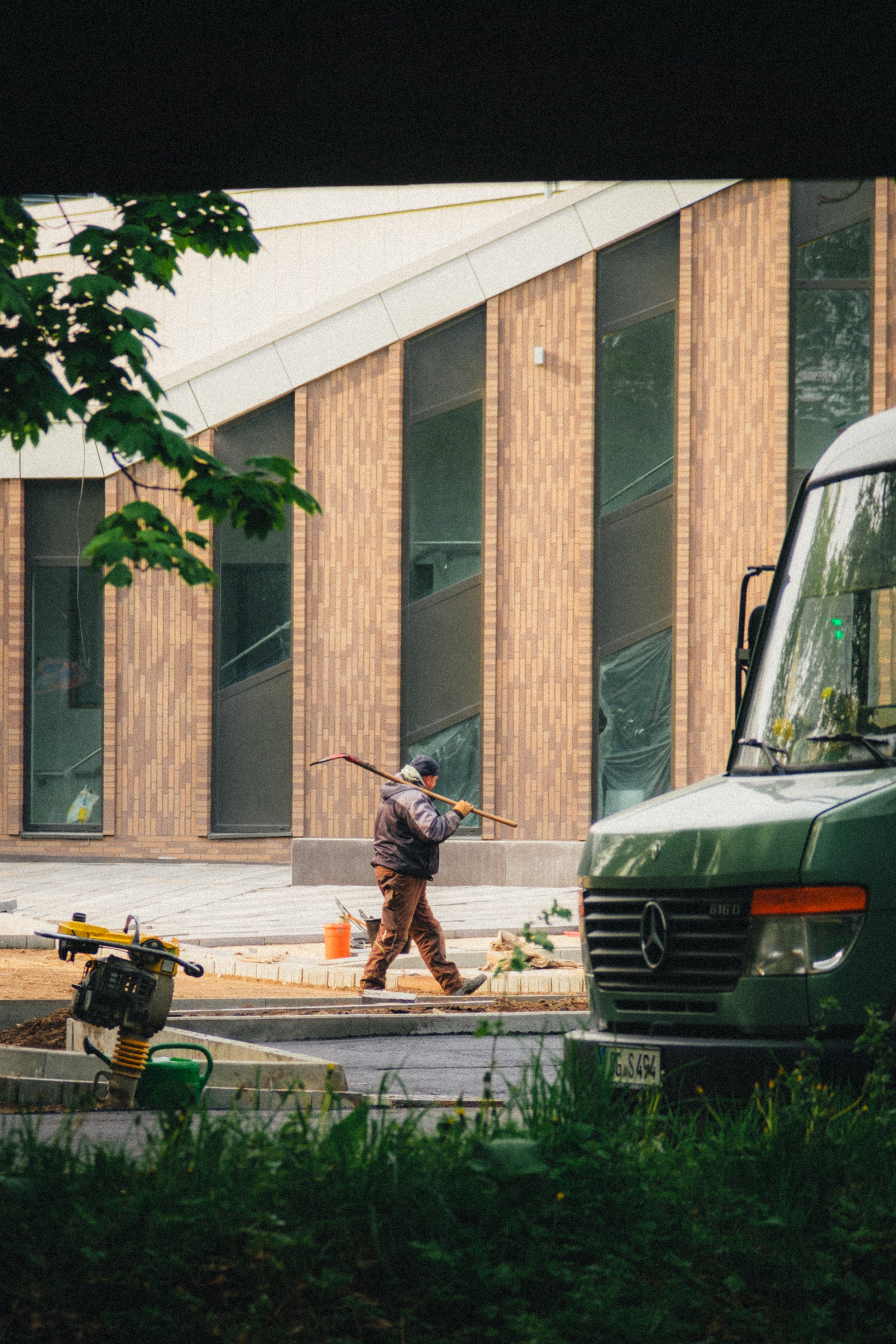 Colorful street photograph of a construction worker carrying tools near a modern brick building in Düsseldorf, Germany. The composition includes a green van and scattered equipment, with natural foliage framing the urban work scene.