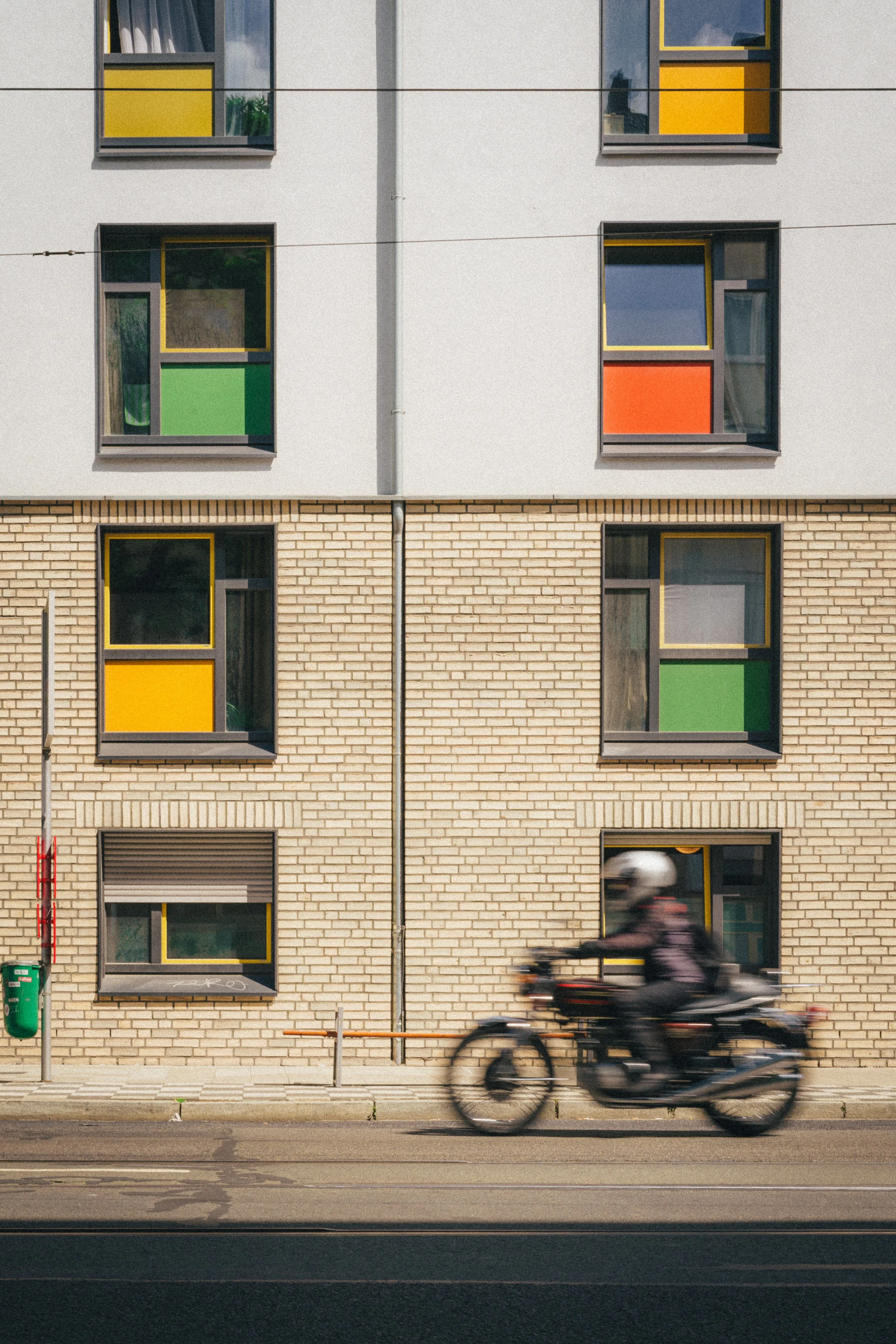 A modern urban scene in Düsseldorf, Germany, featuring a motion-blurred motorcycle passing by a building with colorful geometric windows, showcasing vibrant yellow, green, and red panels against light brickwork.