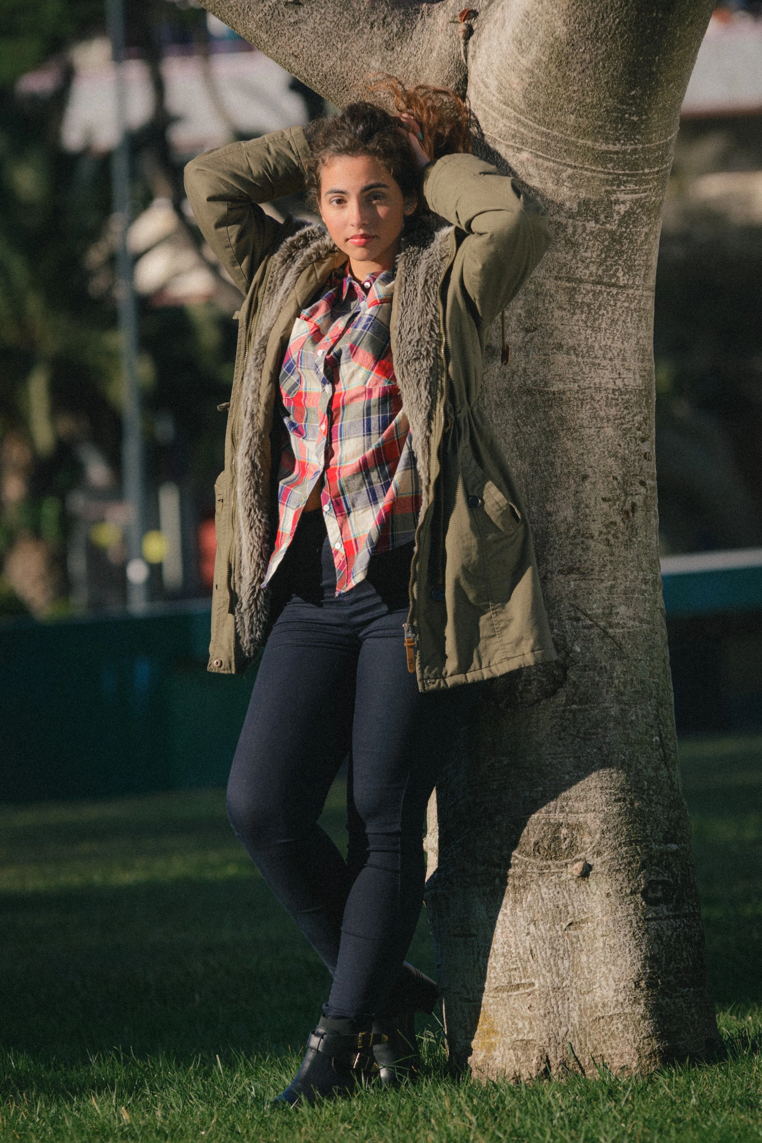 Portrait of Inês Chalupa laying agains a tree, wearing a green coat looking at the camera, grabbing her hair.