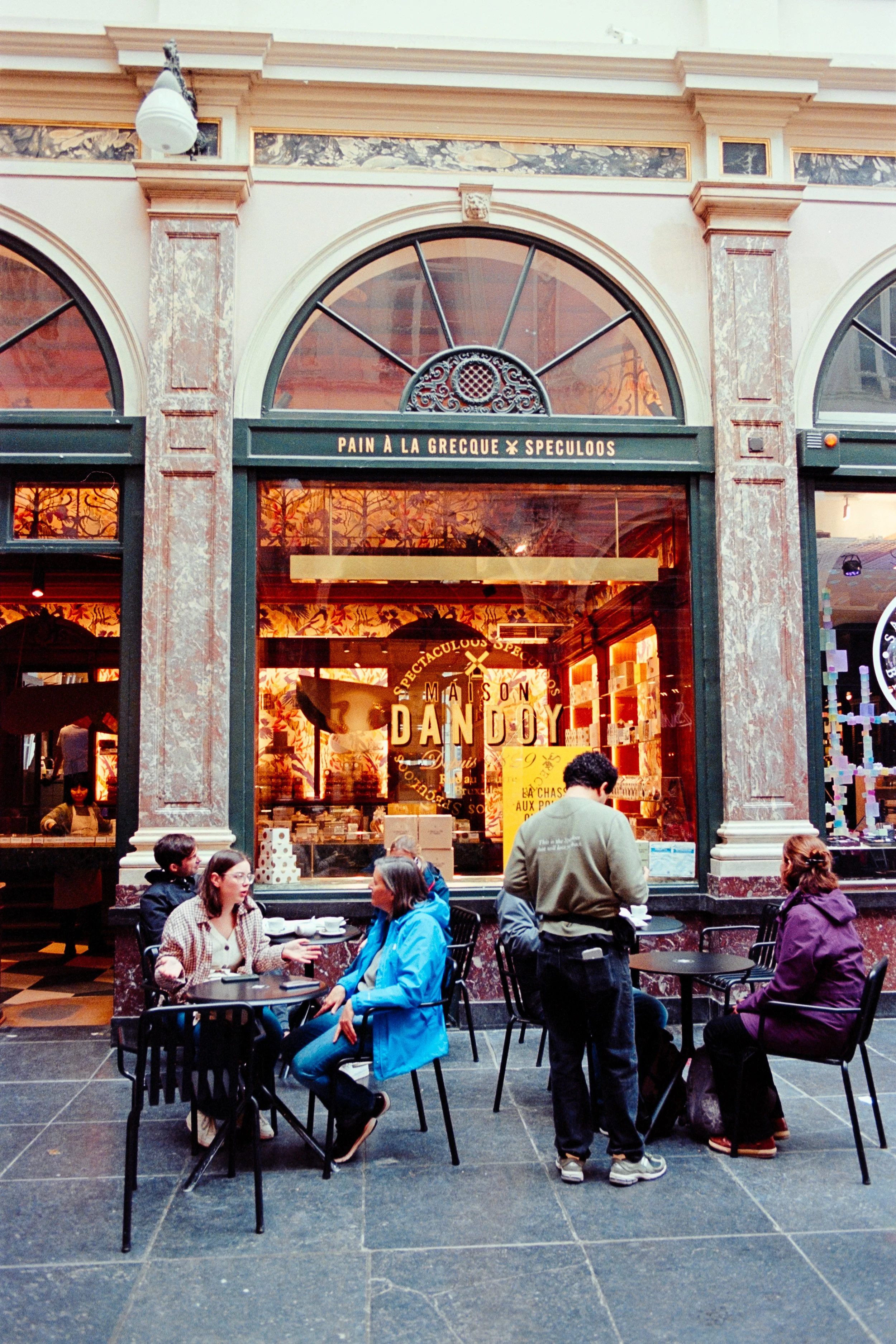 Inside the historic Galeries Royales Saint-Hubert in Brussels, visitors enjoy coffee at a café outside the famous Maison Dandoy store, known for its speculoos biscuits. The elegant gallery features arched windows, ornate designs, and a vibrant atmosp