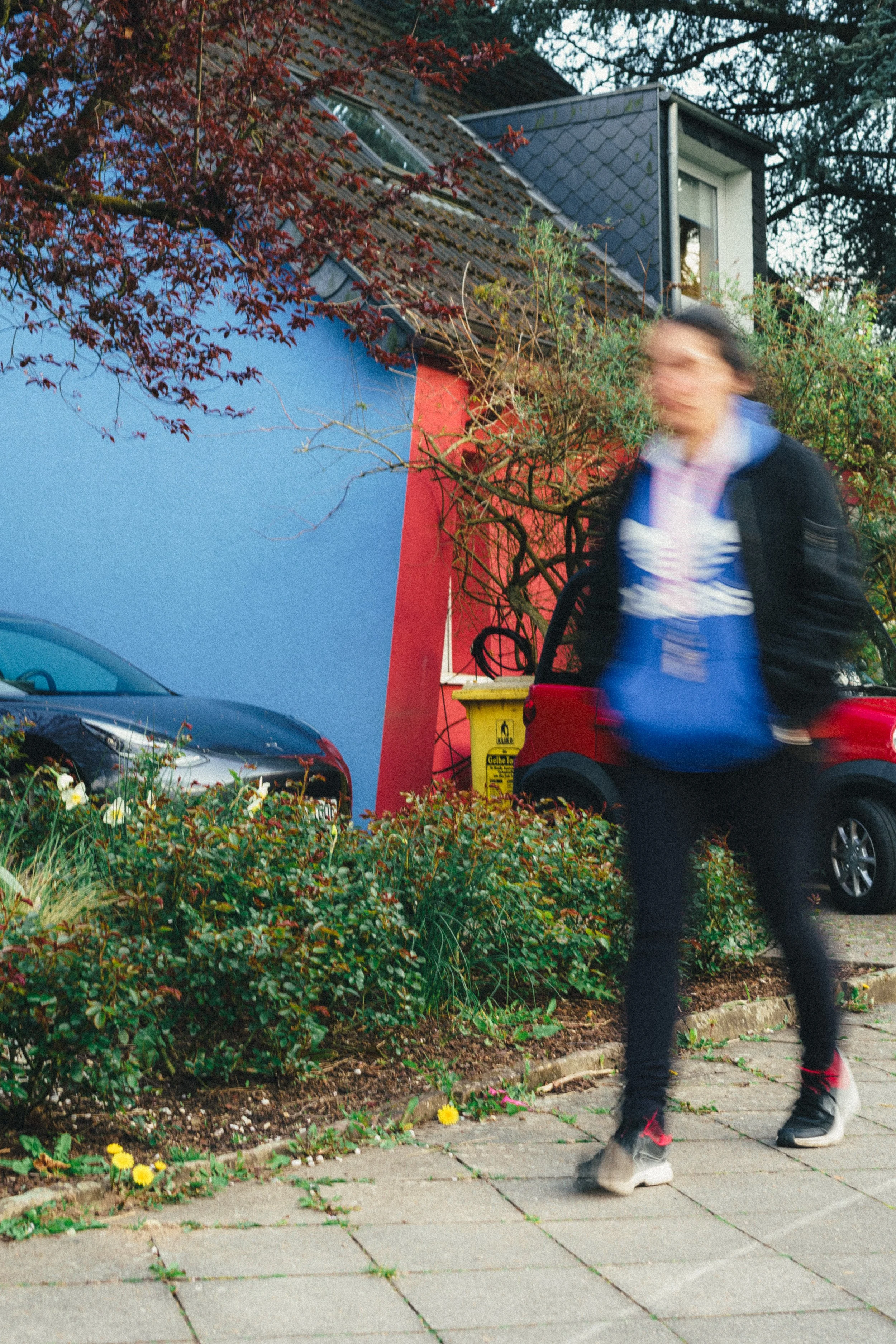 A dynamic street scene in Düsseldorf, Germany, capturing a motion-blurred pedestrian walking past vibrant red and blue house walls, parked cars, and spring foliage.
