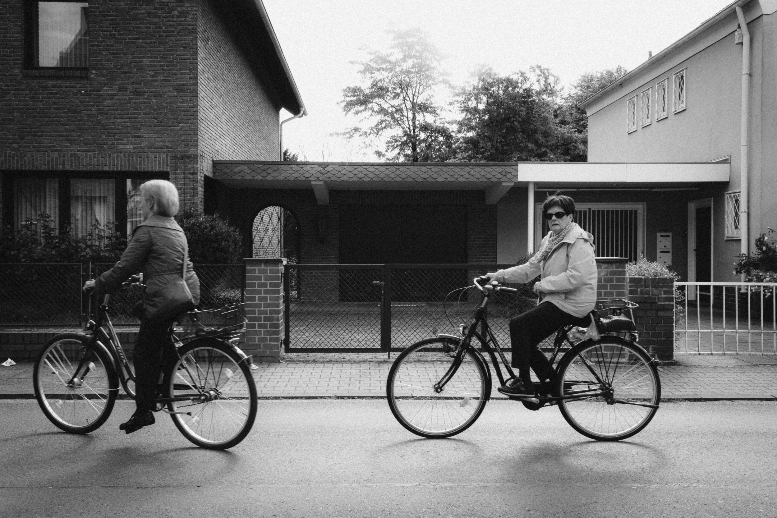 Fine art black and white street photograph of two women cycling past residential houses in Düsseldorf, Germany. The composition captures a moment of everyday life with an urban suburban backdrop, highlighting movement and simplicity.