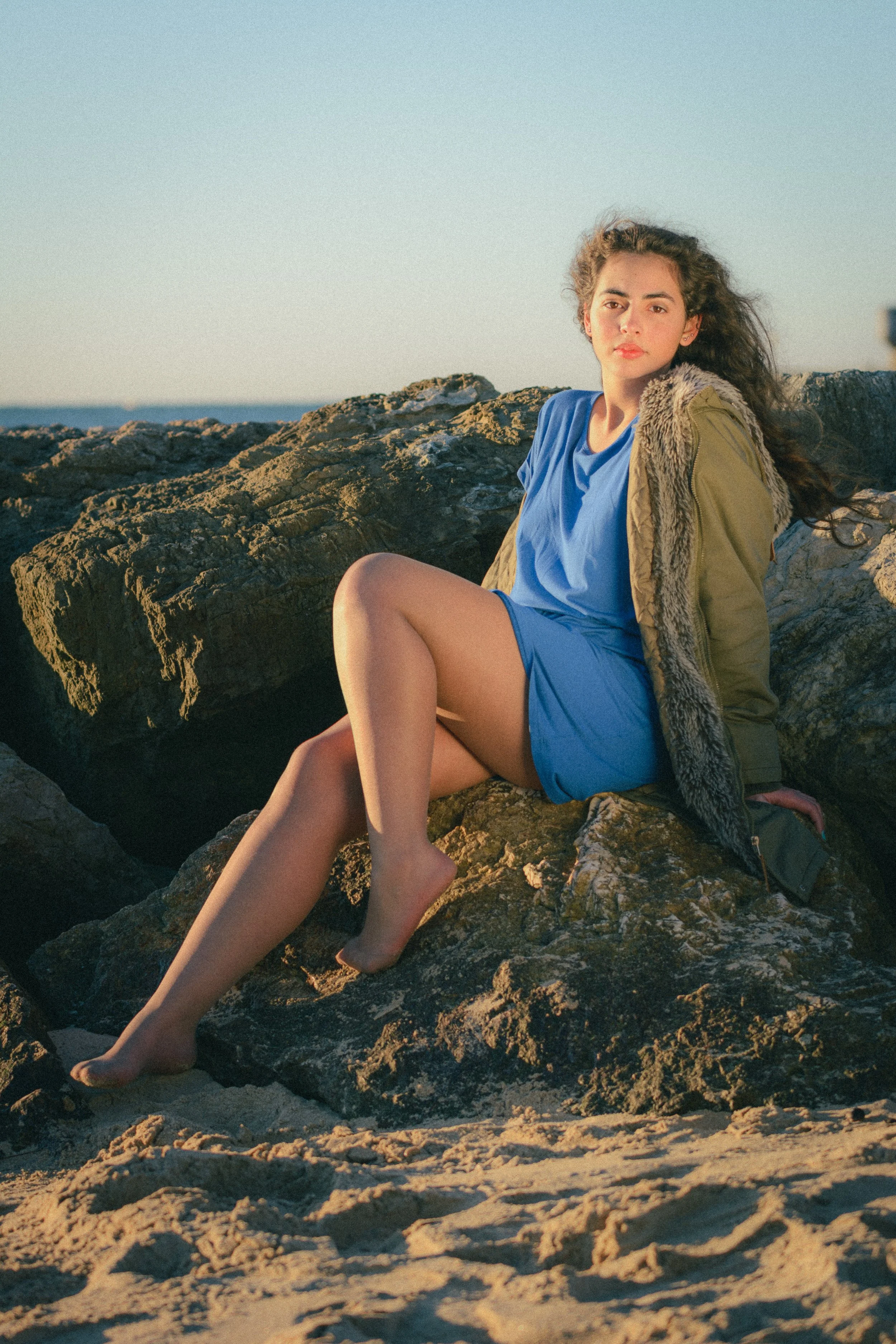 Portrait of Inês Chalupa in a beach in Portugal in Golden hour, wearing a blue dress and a green coat looking at the camera, sitting in the rocks with the blue sky behind her.