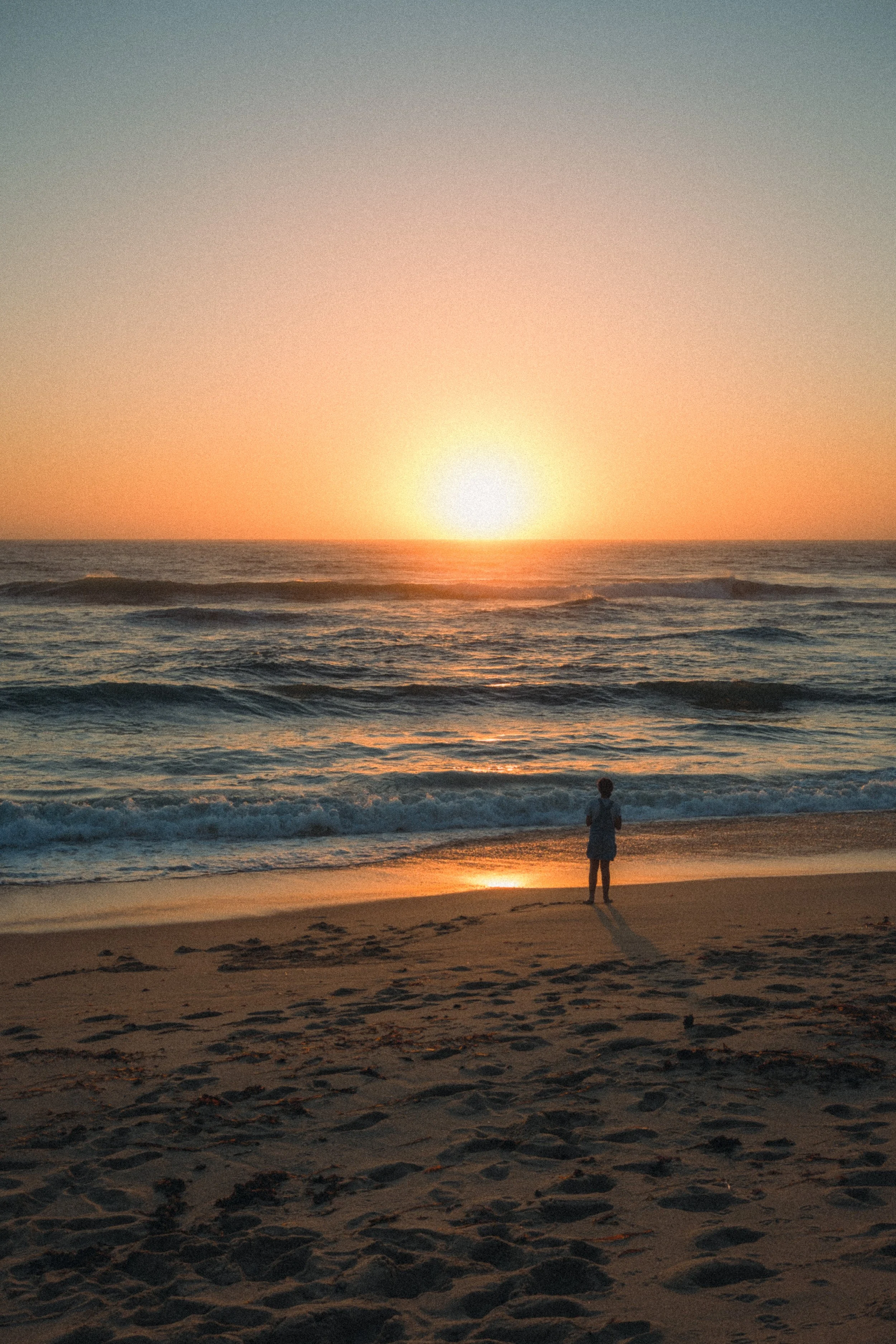 Fine art travel photography of a child standing on the beach in Ericeira, Portugal, gazing at the golden sunset over the ocean waves.