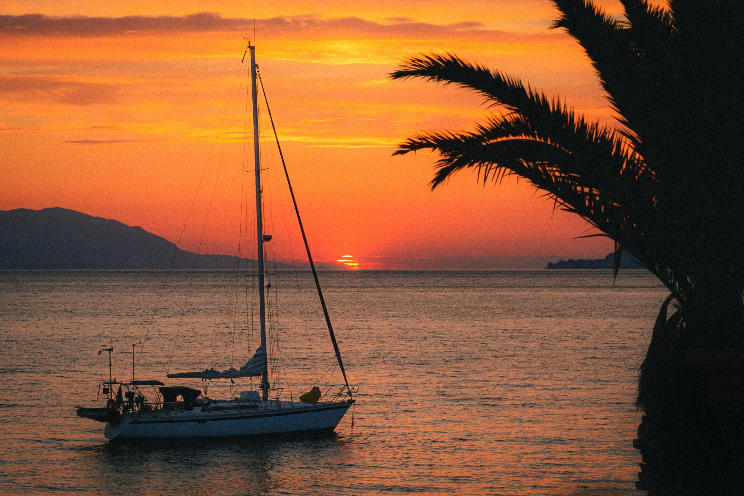 Fine art photograph of a landscape in Loutraki, Greece, featuring a sailing boat against an orange sunset with the sun hiding between the horizon of the sea and the sky. Or the right stands the shadow of a palm tree.