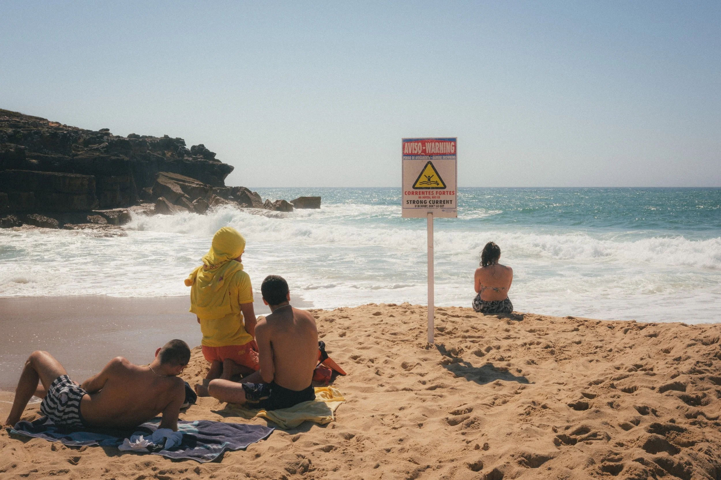 Fine art travel photography of a sunny beach in Ericeira, Portugal, with people lounging on the sand near a strong current warning sign and rocky cliffs by the ocean.