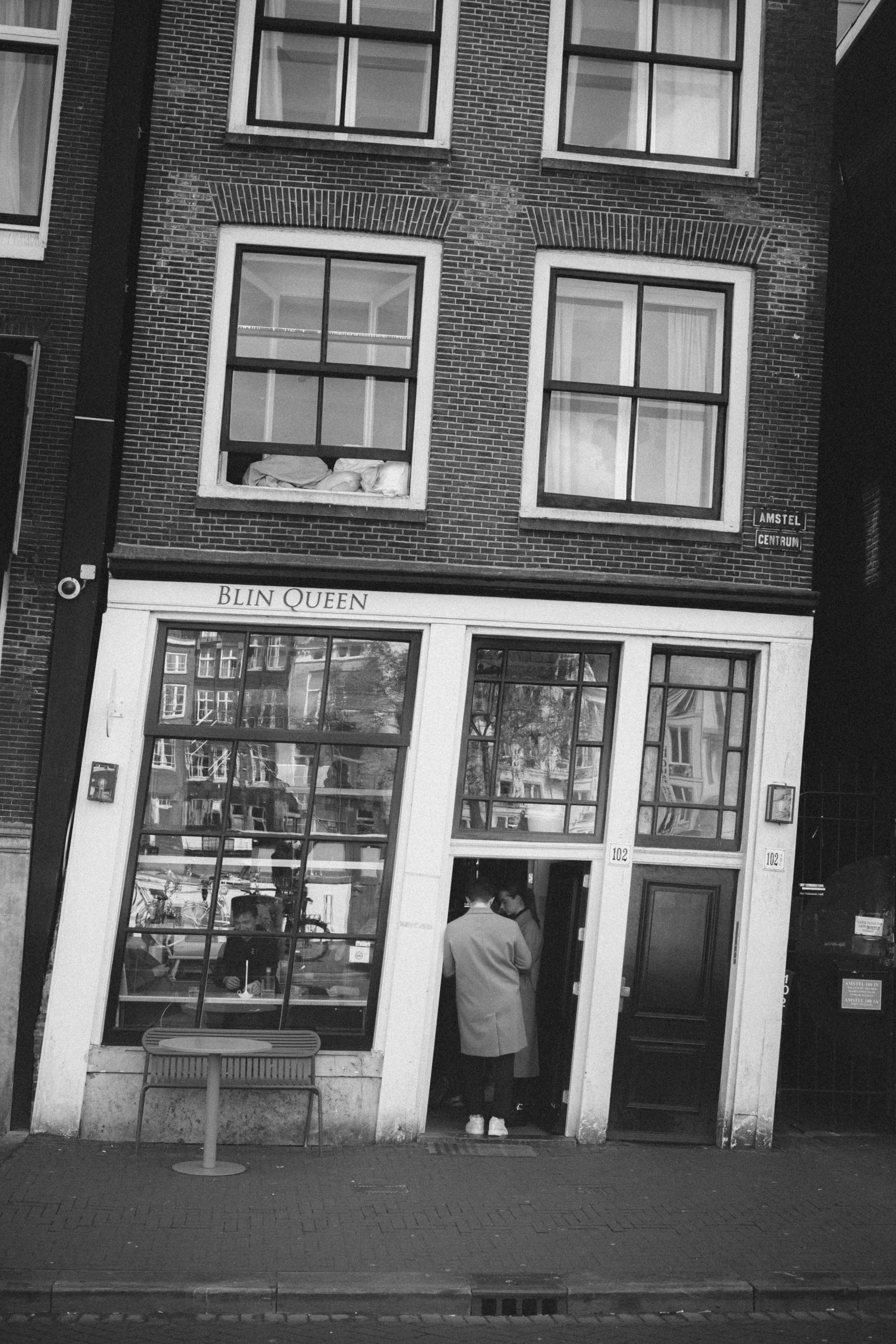 Black-and-white street photography of a leaning building in Amsterdam, Netherlands, featuring large windows reflecting the city's architecture and a cafe named 'Blin Queen' with people interacting at the entrance.