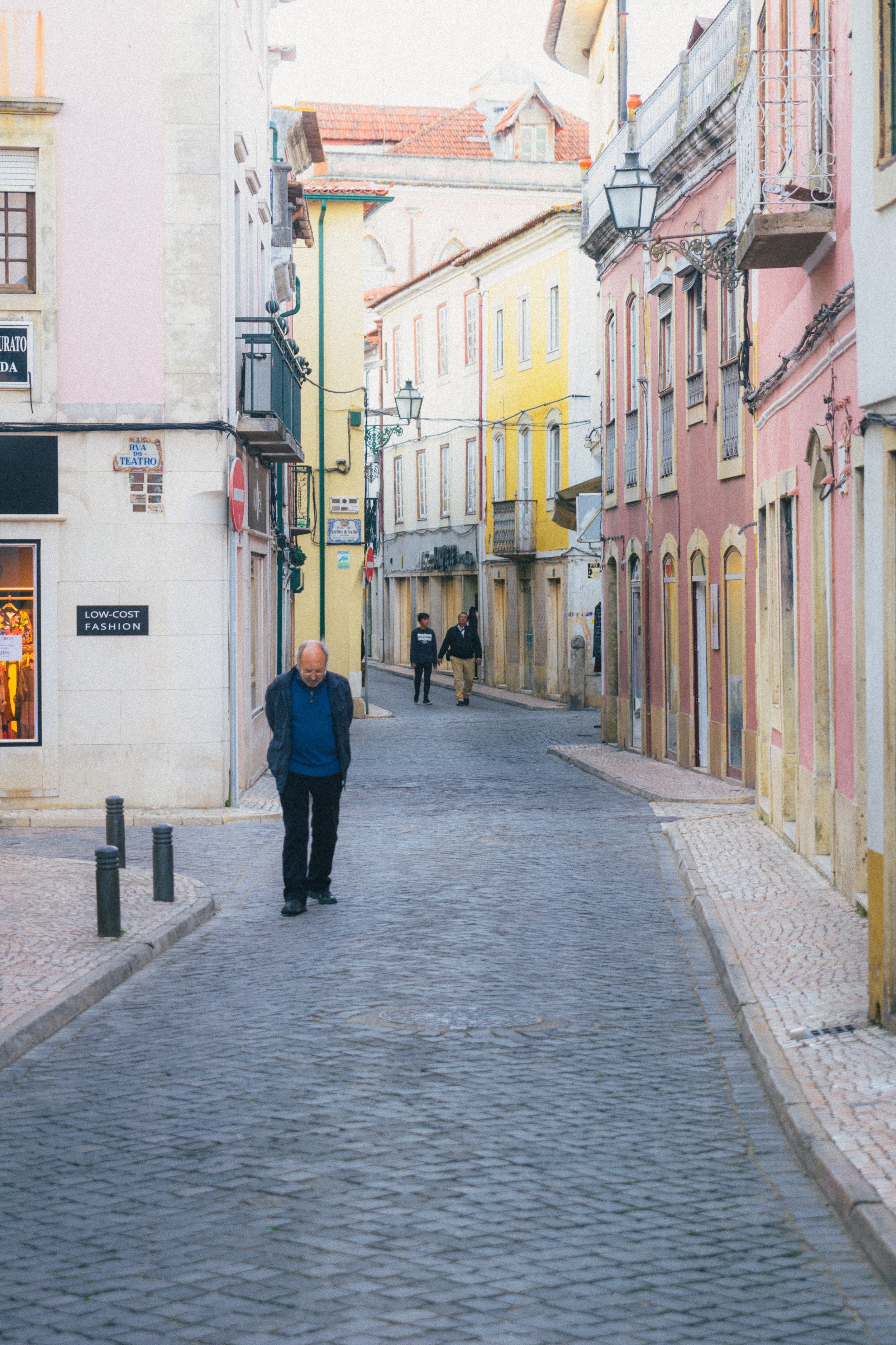 A charming cobblestone street in Tomar, Portugal, flanked by pastel-colored buildings with traditional architectural details. A man walks with his head down, while a few others stroll in the background, evoking a quiet, timeless atmosphere in this hi