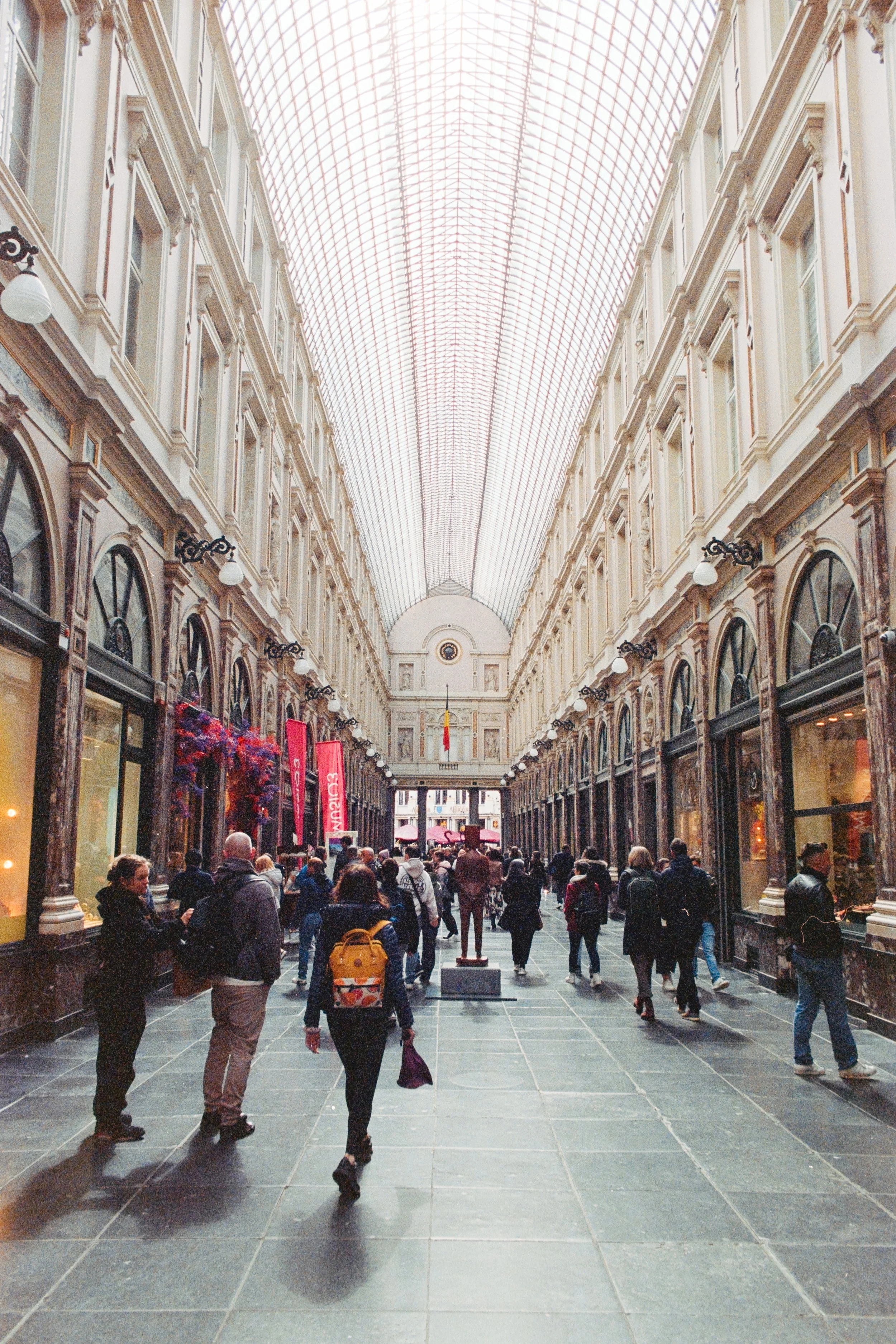 A bustling indoor gallery in Brussels featuring high ceilings with an intricate glass canopy. Elegant storefronts line the passage, and a crowd of people strolls along the polished stone floors, creating a lively atmosphere.