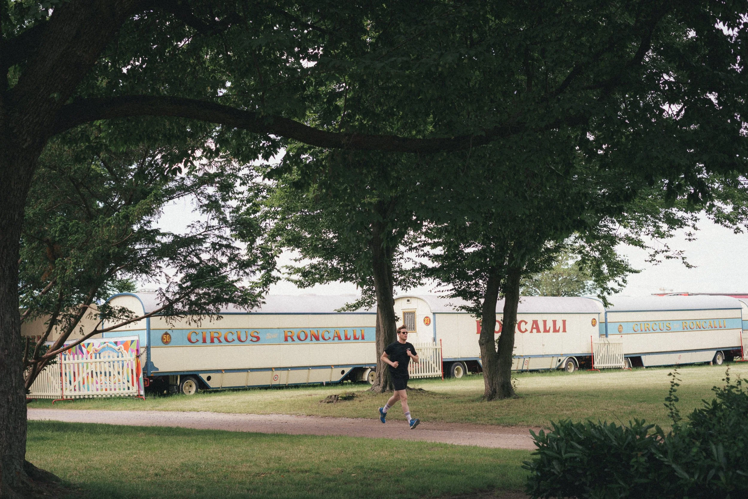 A man jogging along a path in a park framed by large trees, with vintage Circus Roncalli trailers visible in the background. The trailers are painted in cream and blue with red lettering, set on a green grassy field in Düsseldorf.
