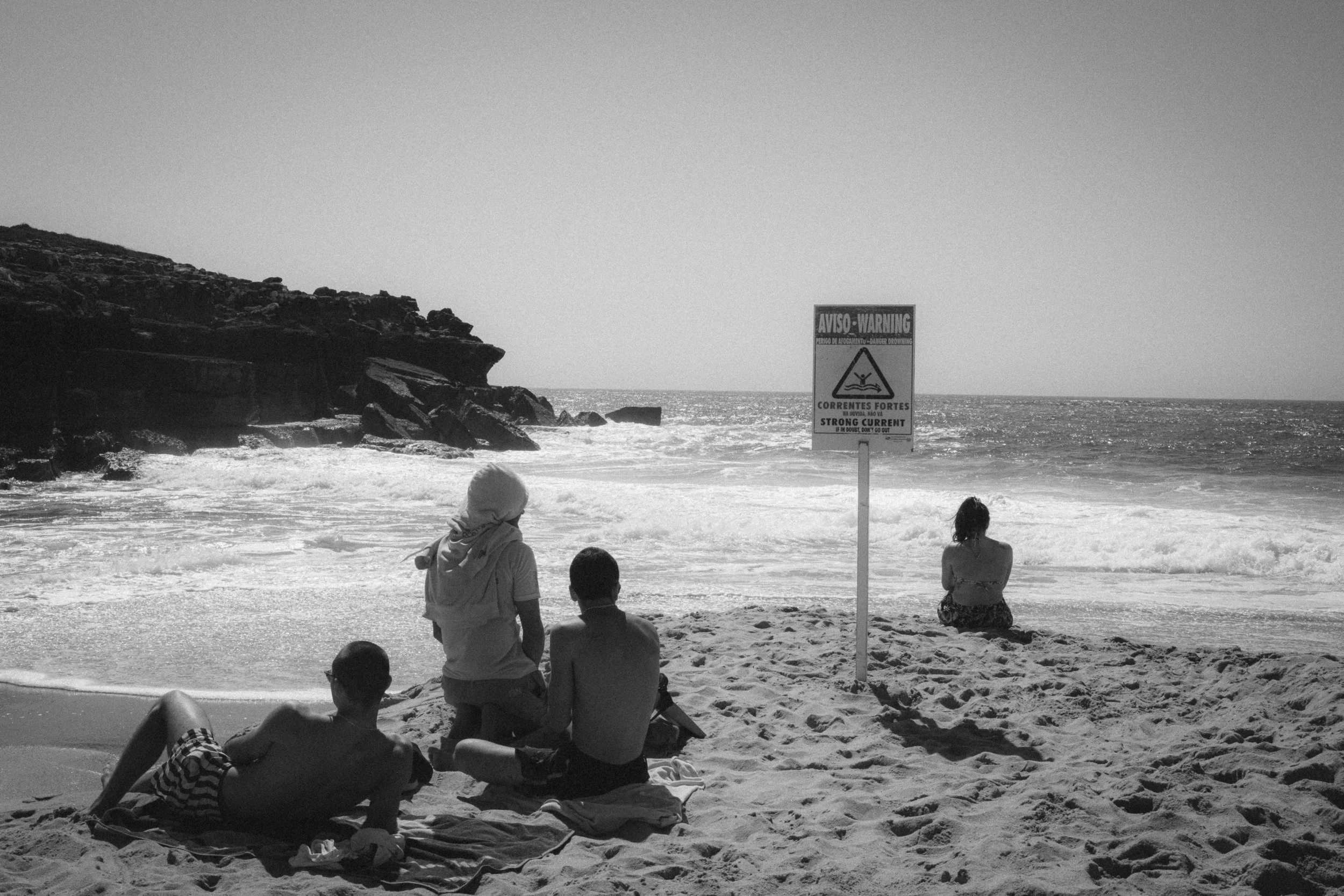 Fine art black-and-white travel photography of a beach in Ericeira, Portugal, with people relaxing on the sand near a warning sign about strong currents and rocky cliffs in the background.