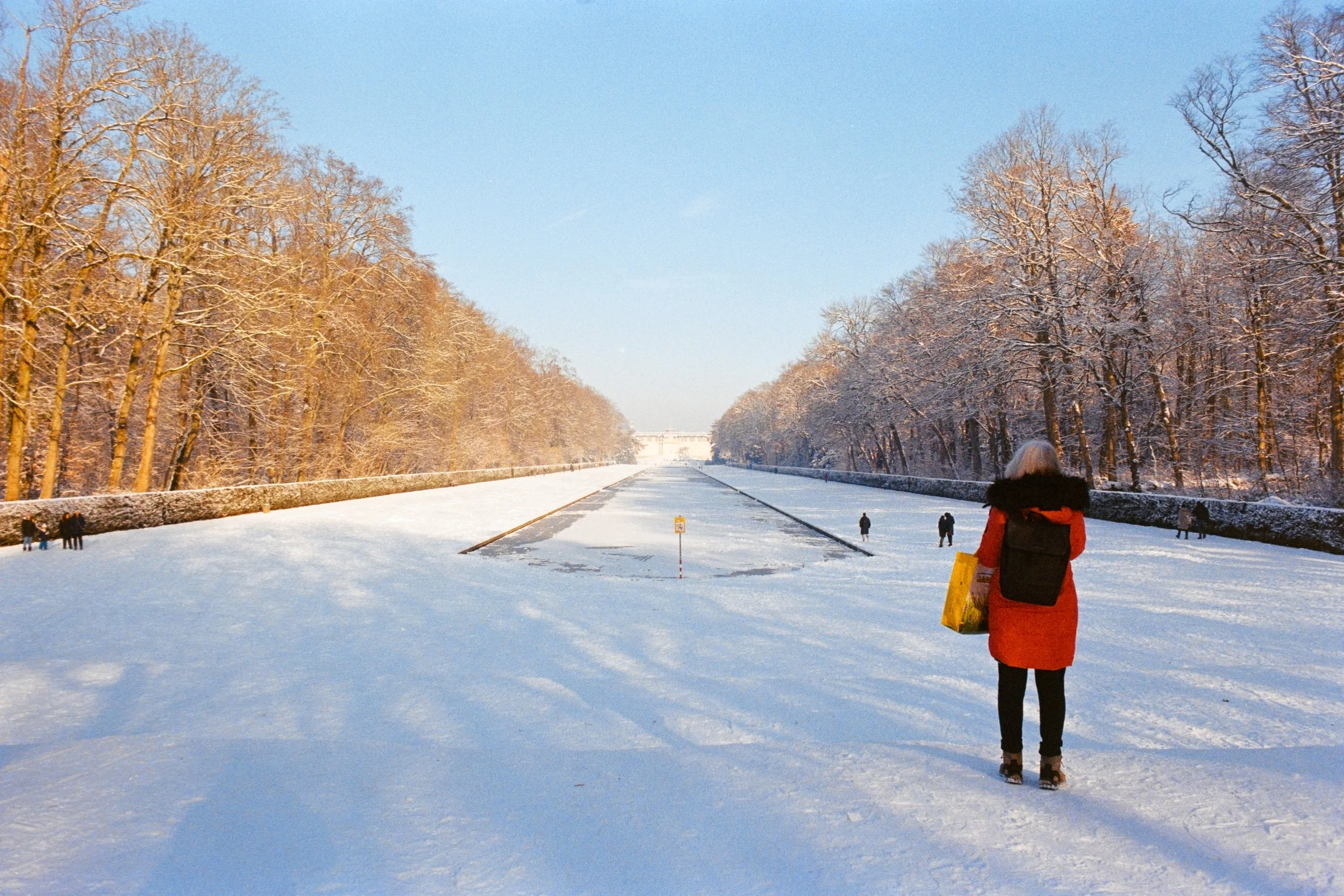 Fine art travel photography of a snow-covered landscape in Schloss Benrath Park, Düsseldorf, Germany, with a person in a red coat standing by a frozen canal framed by trees and a distant view of the palace.