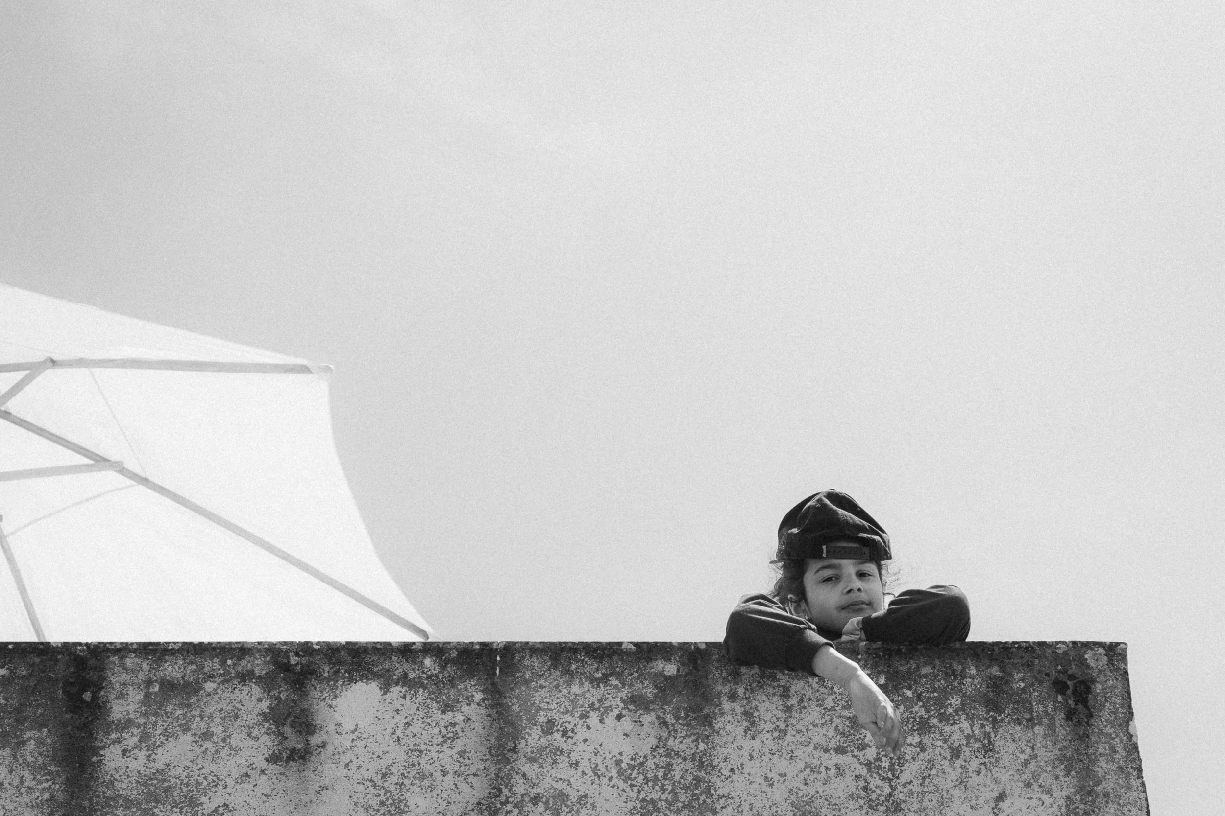 Black and white photograph of Maria Alice Costa leaning over a weathered concrete wall in Ericeira, Portugal. The minimalistic composition features a bright umbrella in the background and a serene sky, emphasizing her thoughtful expression.