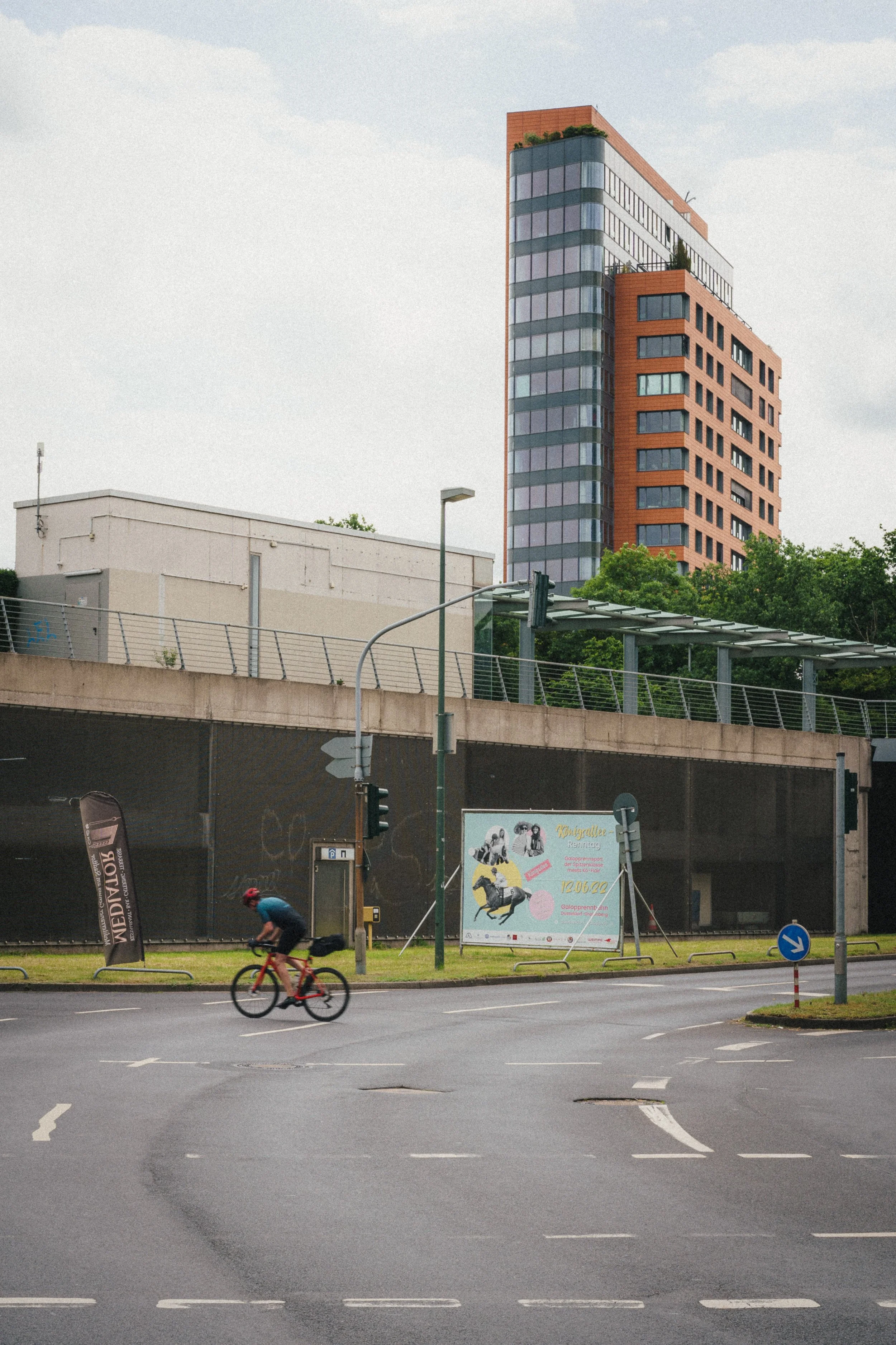 A cyclist wearing a red helmet and blue shirt rides across a wet intersection in Düsseldorf. In the background, a modern orange and gray high-rise building towers above an overpass with greenery and advertisements.