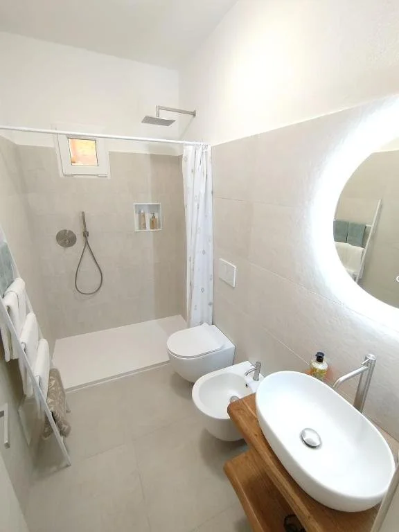 Modern bathroom with walk-in shower, toilet, bidet, oval sink on a wooden countertop, round mirror, and towel rack.