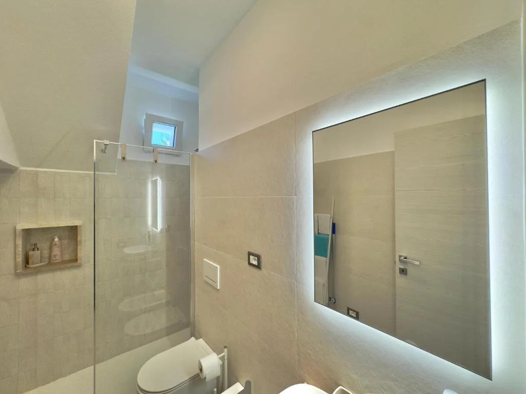 Modern bathroom with a large backlit mirror, toilet, and walk-in shower with a small window.