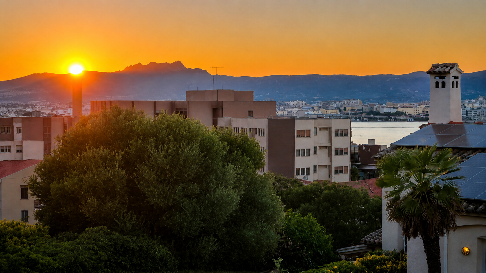 Sunset over a city with mountains in the background, residential buildings, greenery, including a palm tree, and solar panels on a roof.