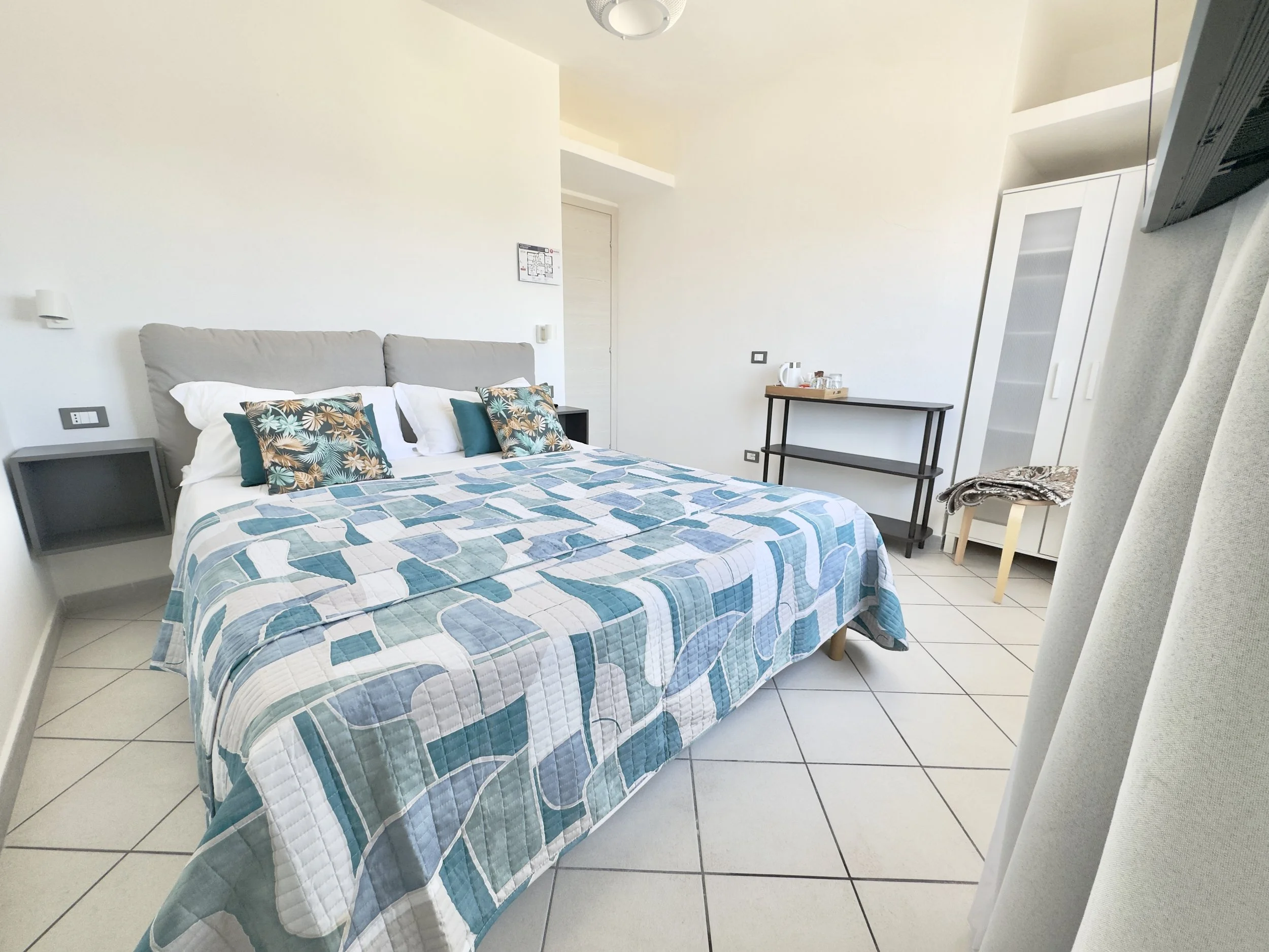 A hotel room with a bed featuring a blue and white patterned quilt and tropical-themed pillows, a small bedside table, a wooden luggage rack with a tea kettle and cups, a white wardrobe, and a chair with a blanket draped over it.