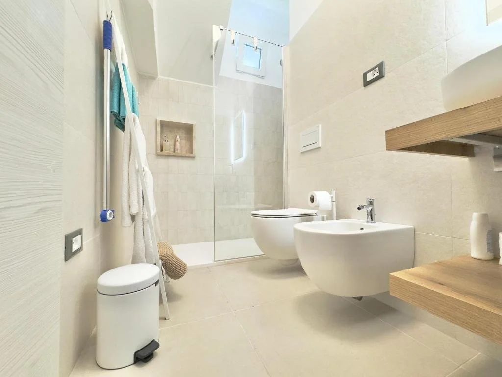 A modern bathroom with a walk-in shower, a toilet, a bidet, and a wooden shelf on the right. The bathroom has beige tiles and a small window near the ceiling.