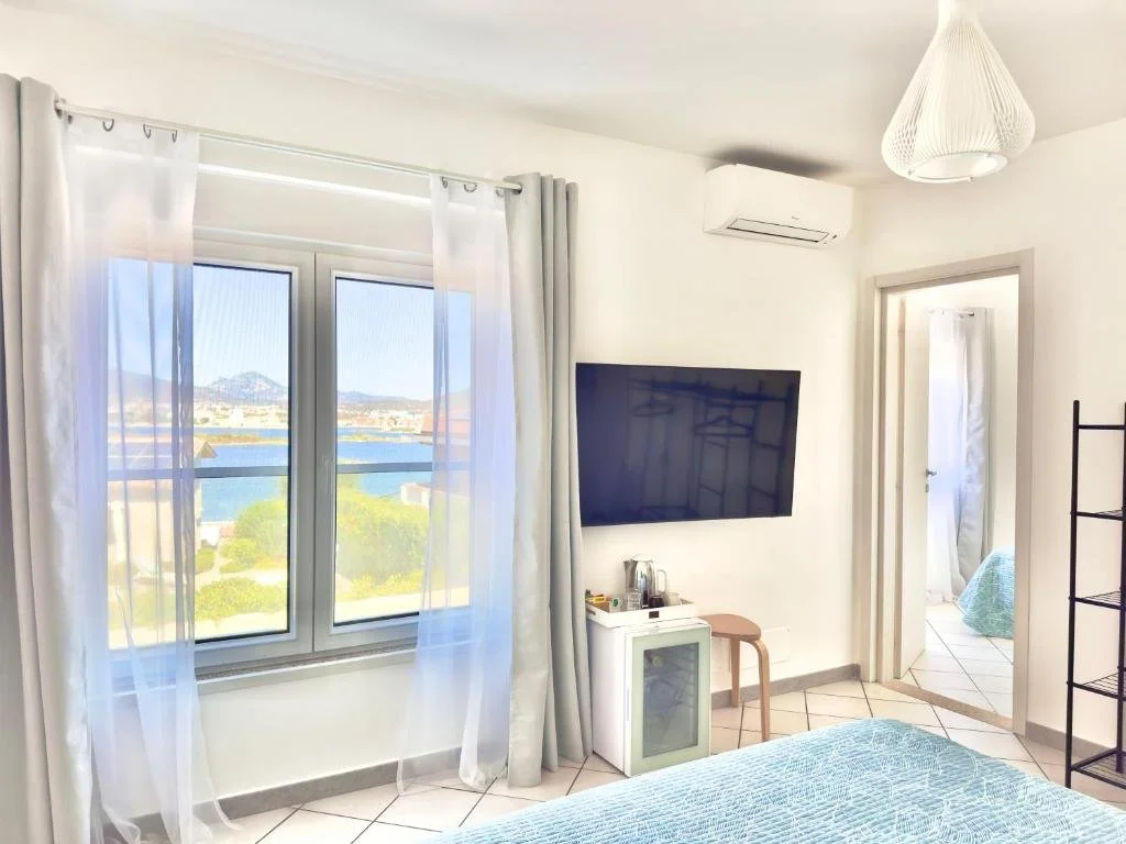 Cozy bedroom with a large window showing a lake and mountains, sheer curtains, wall-mounted TV, small minibar, and an indirect view of an adjacent room with a bed.